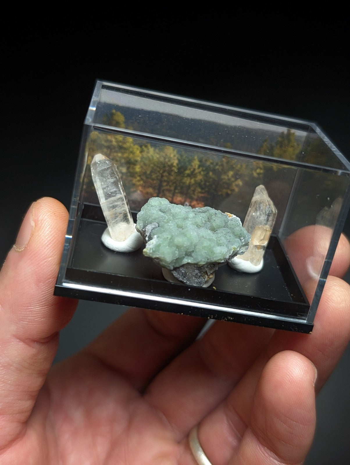 A clear acrylic display case holding a central green mineral specimen flanked by two transparent quartz crystals on white bases.