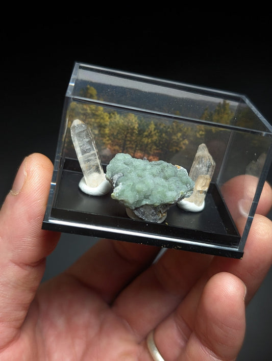 A clear acrylic display case holding a central green mineral specimen flanked by two transparent quartz crystals on white bases.