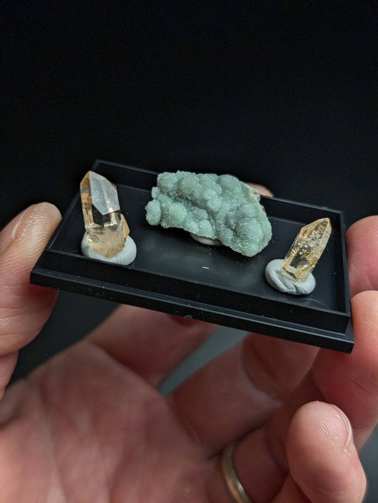 Black rectangular display tray holding three mineral specimens: two golden-yellow quartz crystals with clear facets and a light green, textured rock formation.