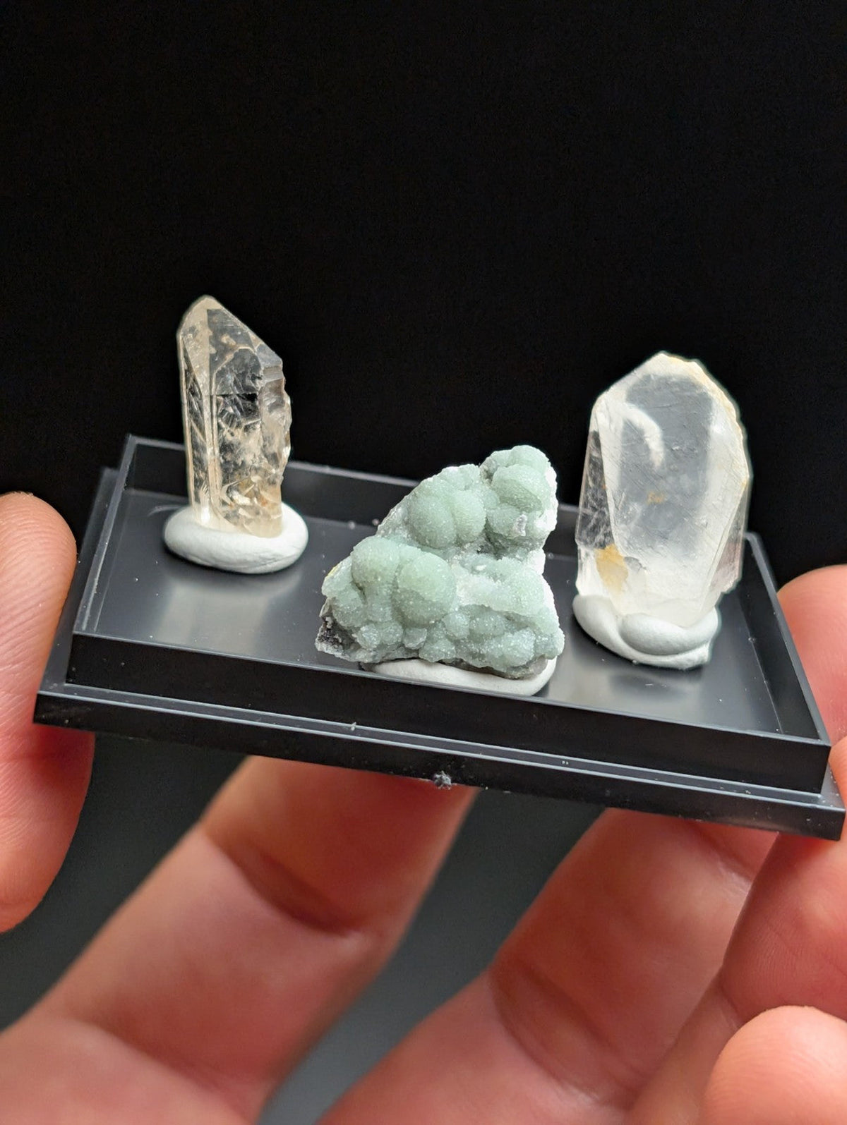 A black display tray holding three distinct mineral specimens: two transparent quartz crystals with internal inclusions and a central cluster of pale green, textured minerals.