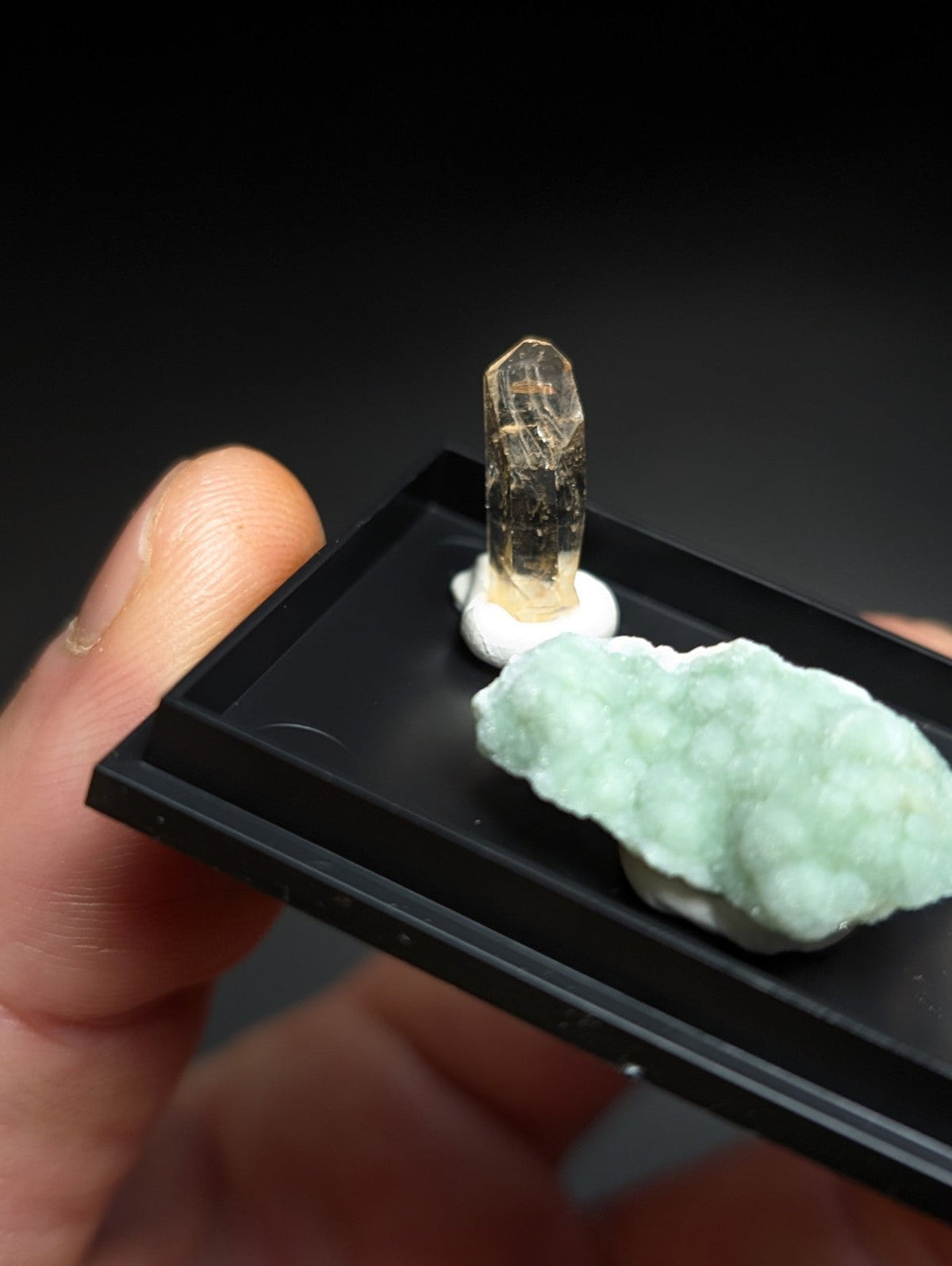 A translucent golden-yellow crystal with a pointed tip, mounted on a white base alongside a light green, bumpy mineral specimen.