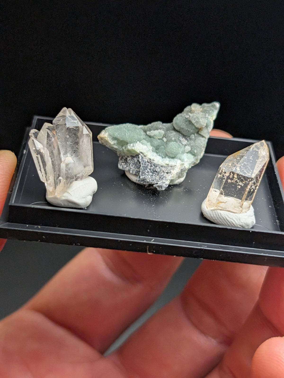 A black display tray holding three distinct mineral specimens: a clear quartz cluster, a greenish-gray geode with white crystalline formations, and another transparent crystal on a white base.