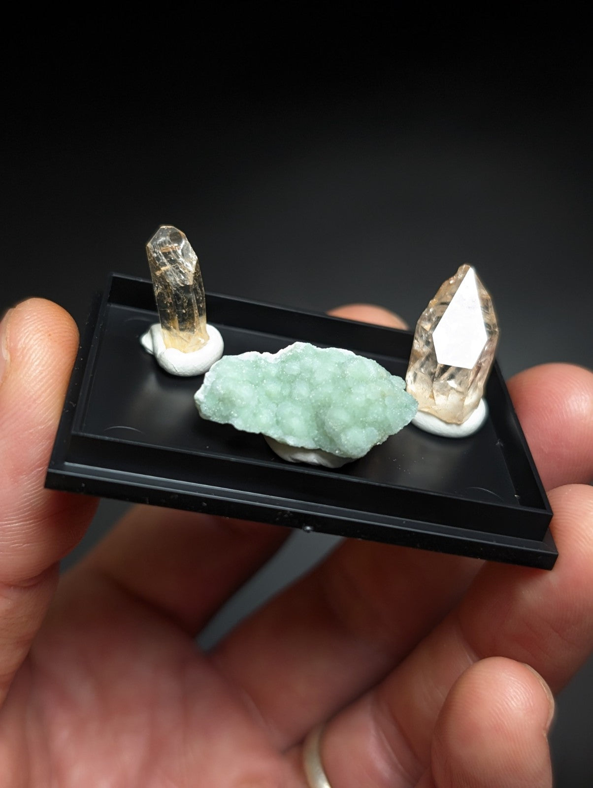 A black display tray holding two clear quartz crystals and a light green mineral specimen.