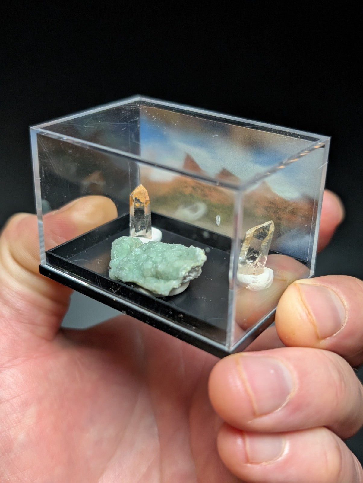 A clear acrylic display case holding a green mineral specimen and two quartz crystals on white stands.