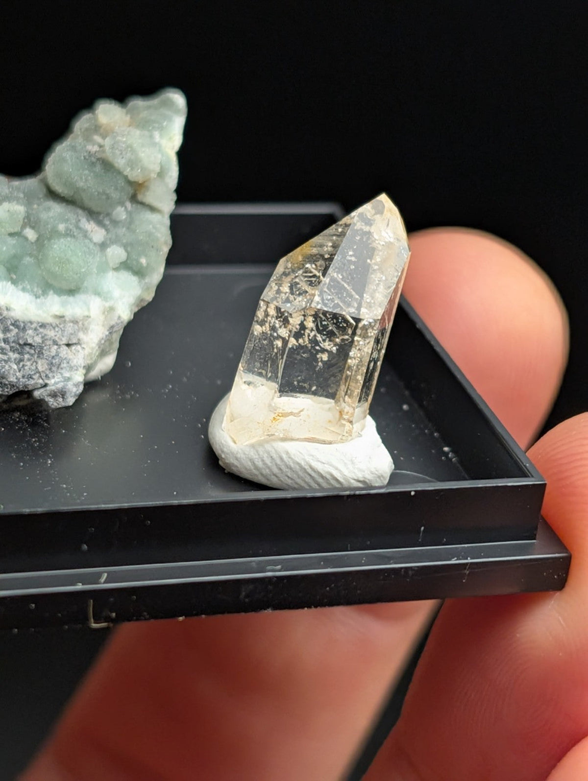 A clear, faceted quartz crystal with subtle golden inclusions sits atop a white, textured base within a black display tray.