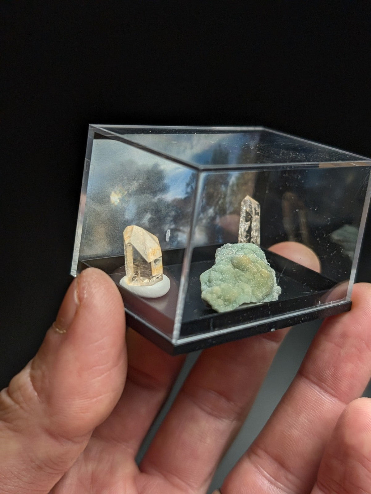 A clear acrylic display case holding two miniature mineral specimens, one with a pale yellowish crystal and the other a greenish rock.
