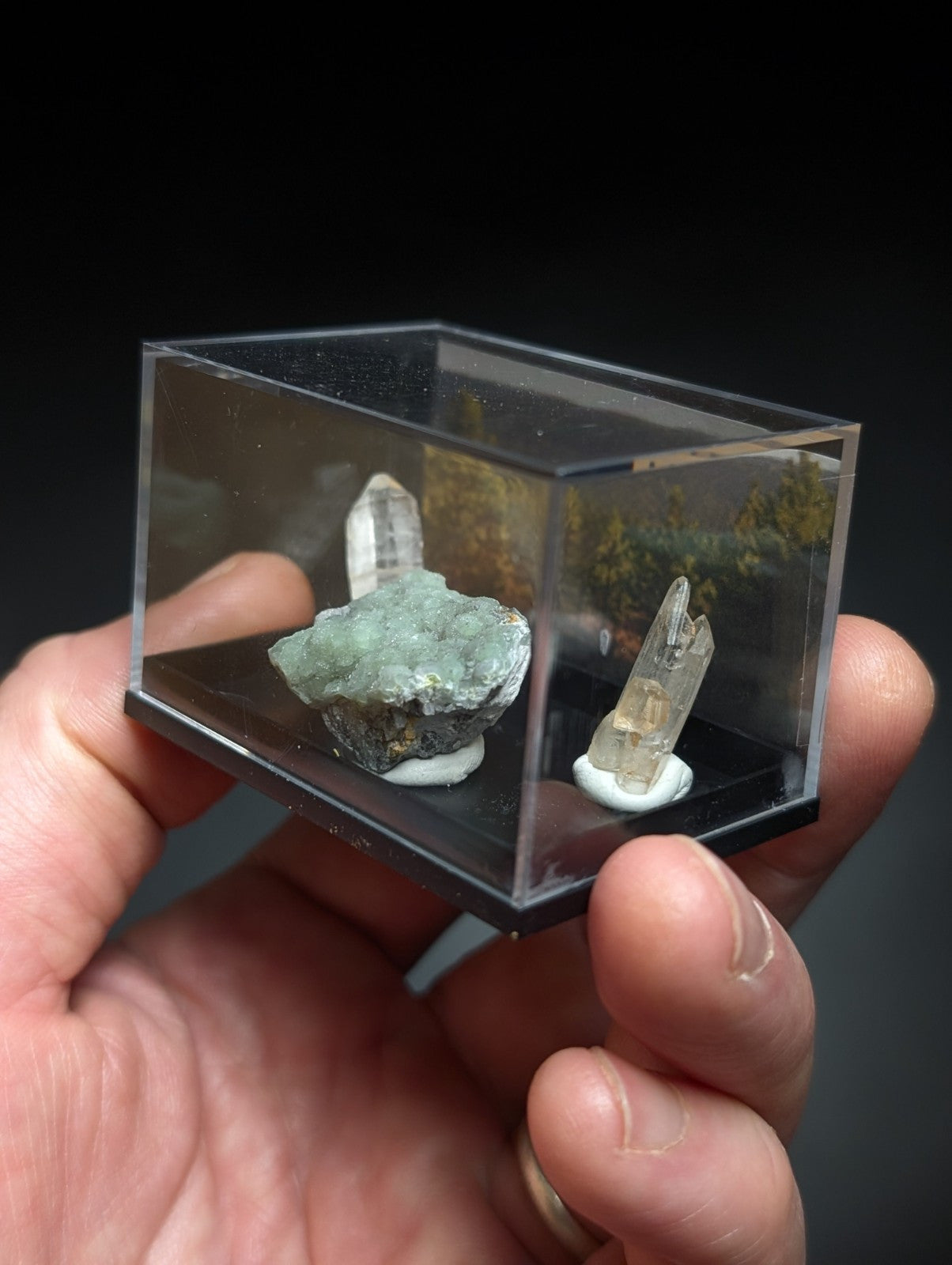 A clear acrylic display case holding two mineral specimens, one with a greenish-blue crystalline formation and another featuring translucent quartz crystals.