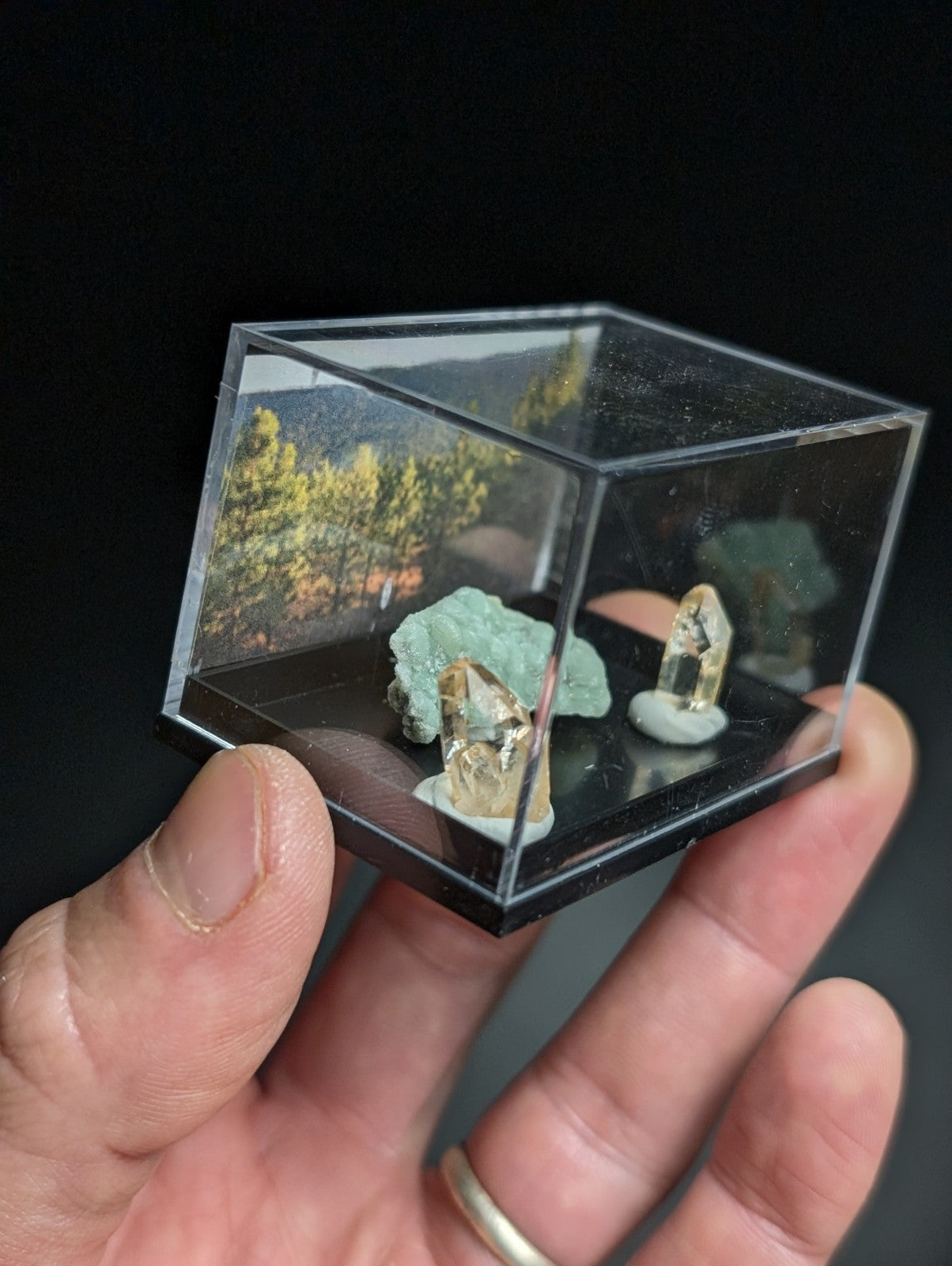 A clear acrylic cube display case holding miniature mineral specimens, including a greenish-blue crystal cluster and two translucent yellow crystals on white bases.
