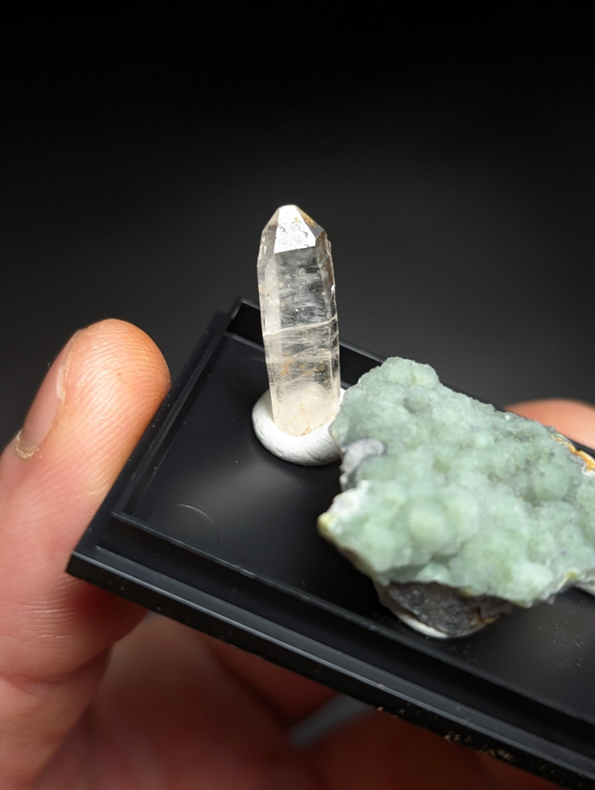 A clear, faceted quartz crystal stands upright on a black display tray beside a textured green mineral specimen.