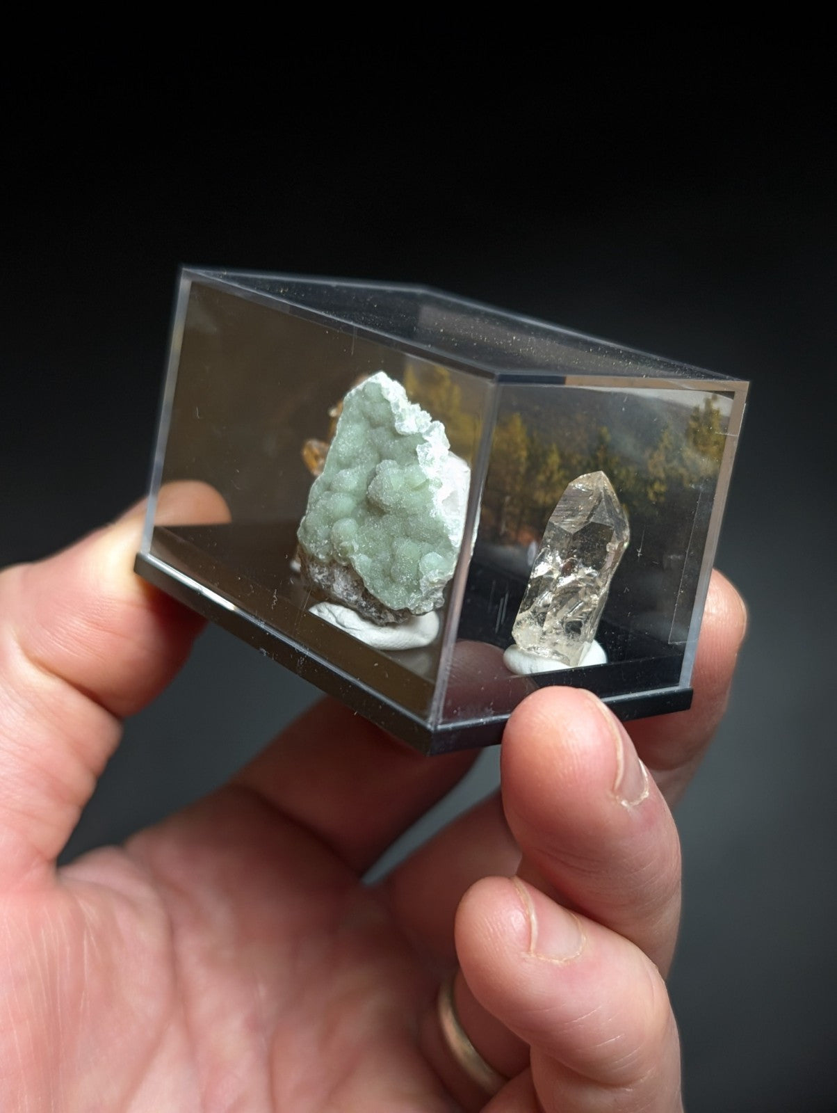 A clear acrylic display case holding two mineral specimens: a greenish-blue crystalline formation and a transparent quartz crystal.