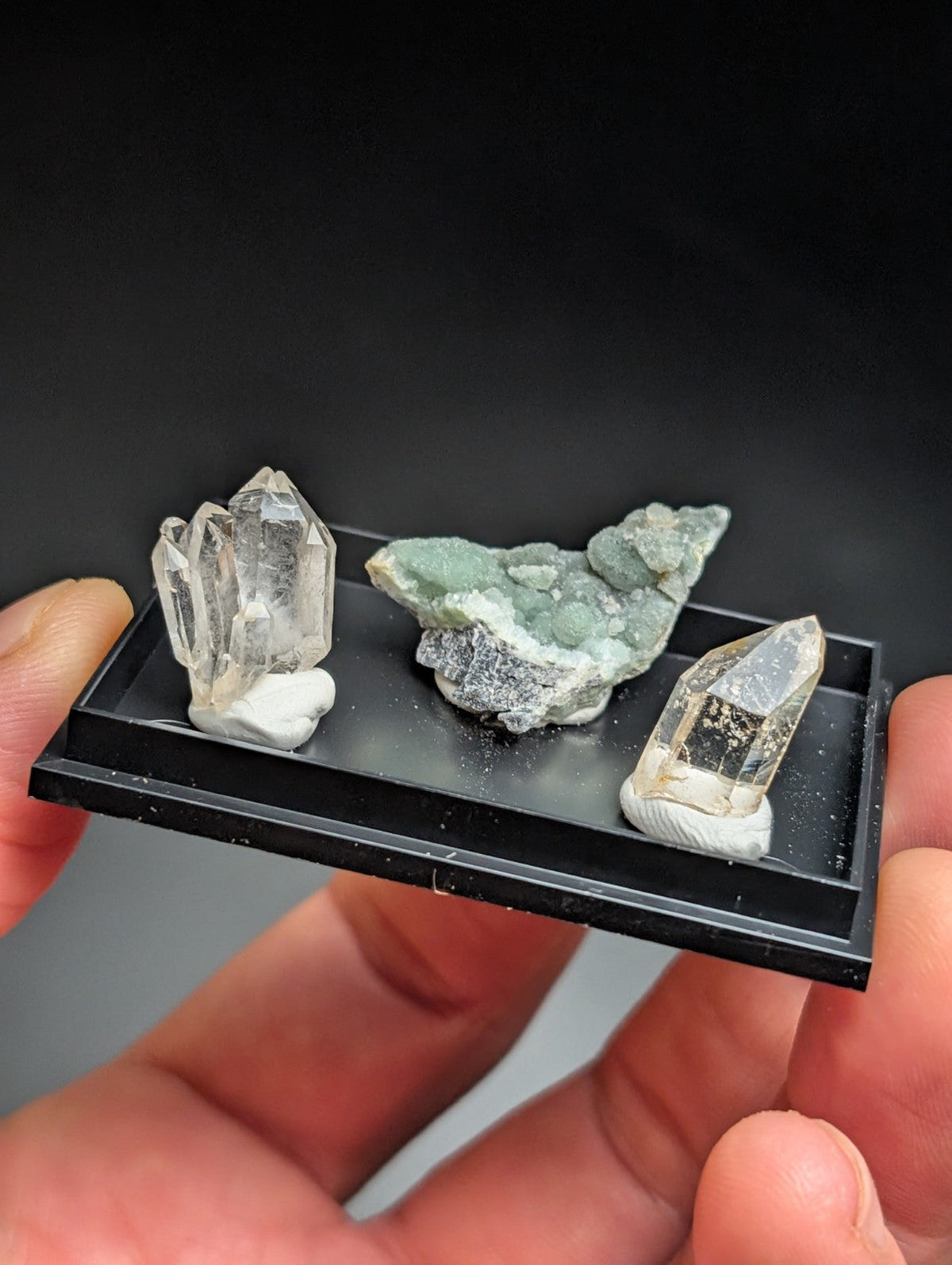 A black rectangular display tray holding three distinct mineral specimens: a clear quartz cluster, a greenish-gray rock formation with crystalline textures, and another transparent crystal on a white base.