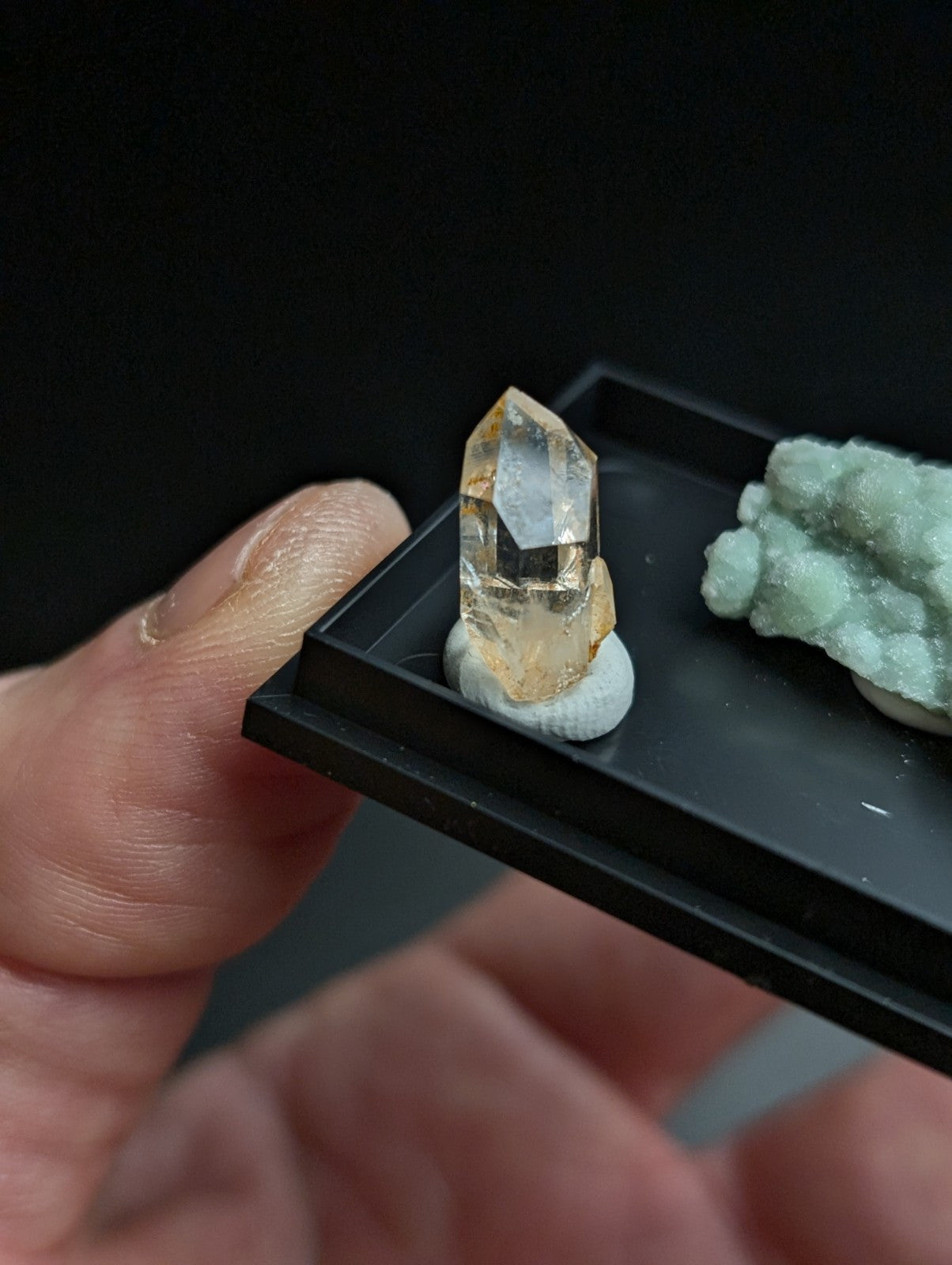 A translucent golden-yellow crystal with a pointed, faceted top and rougher base sits on a small white pedestal within a black display tray.