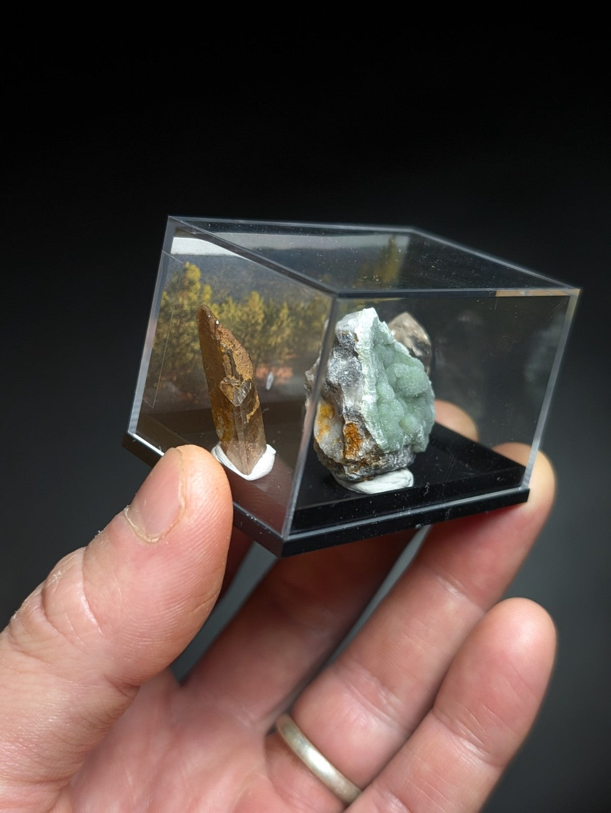 A clear acrylic display case holding two mineral specimens, one with a brownish-orange crystalline formation and another featuring greenish-blue crystals embedded in rock.