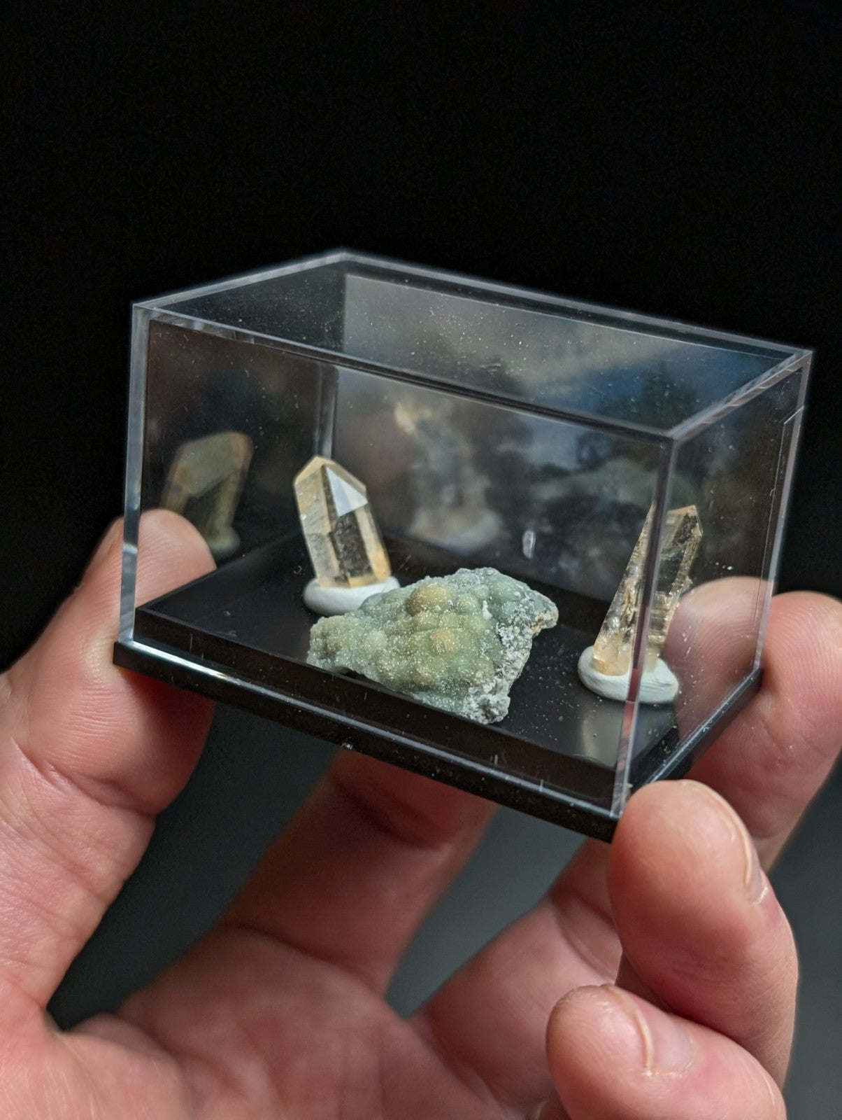 A clear acrylic display case holding two golden-yellow quartz crystals and a greenish mineral specimen on a black base.