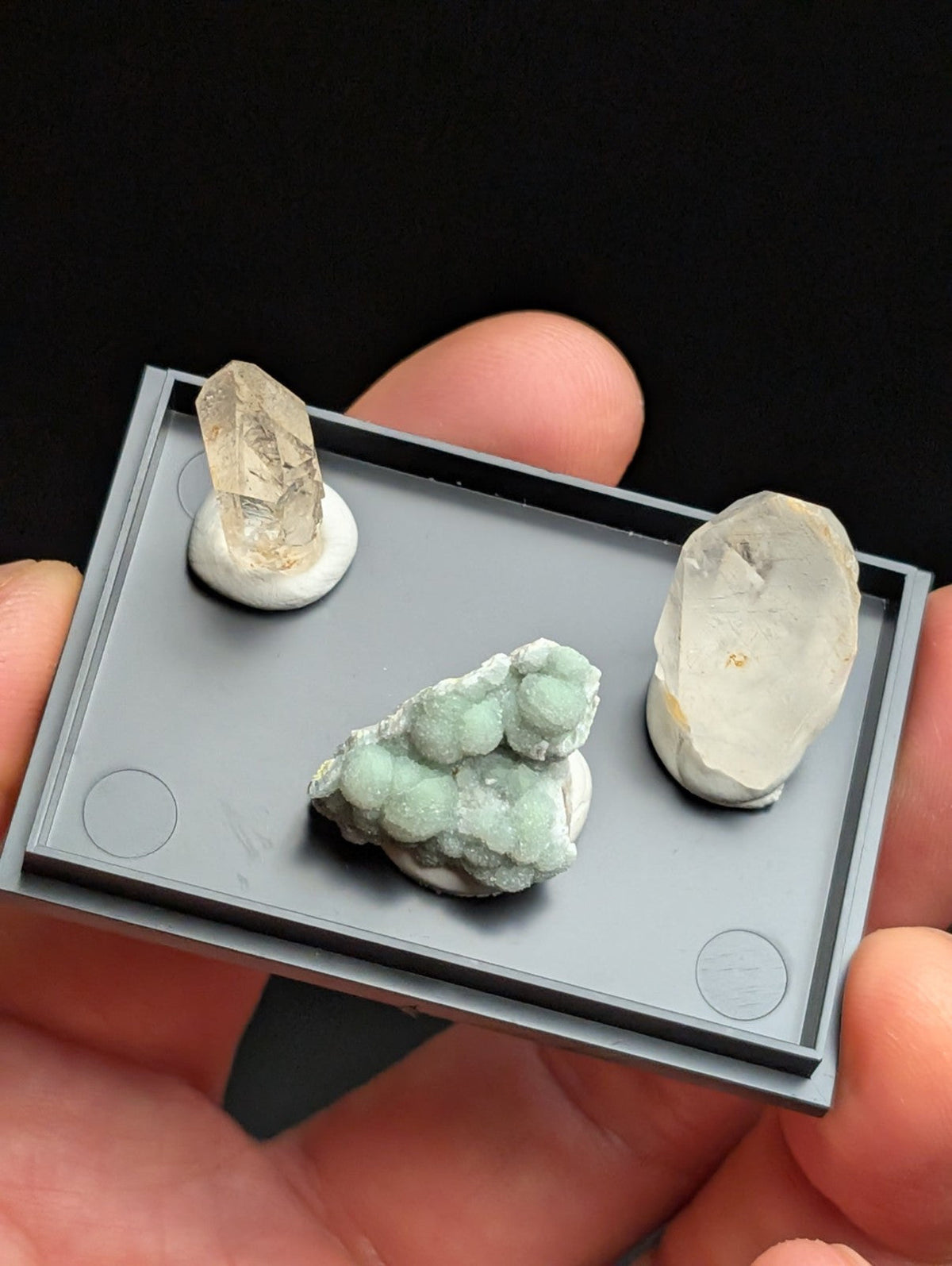 Gray rectangular mineral display tray holding three distinct specimens: a translucent yellowish crystal, an opaque white quartz-like stone with visible internal inclusions, and a cluster of light green crystalline formations.
