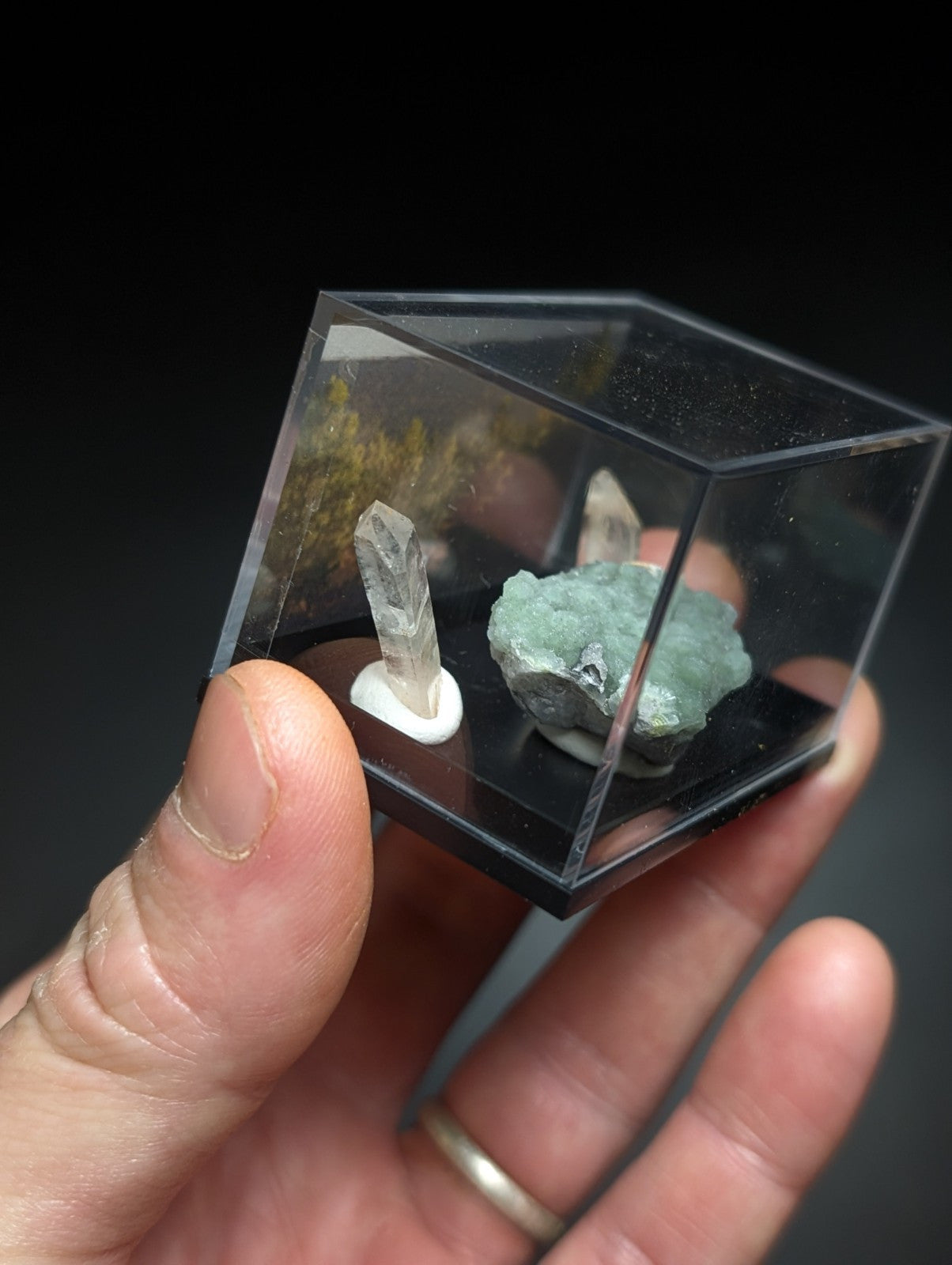 A clear acrylic cube display case holding a transparent quartz crystal and a greenish-blue mineral specimen.
