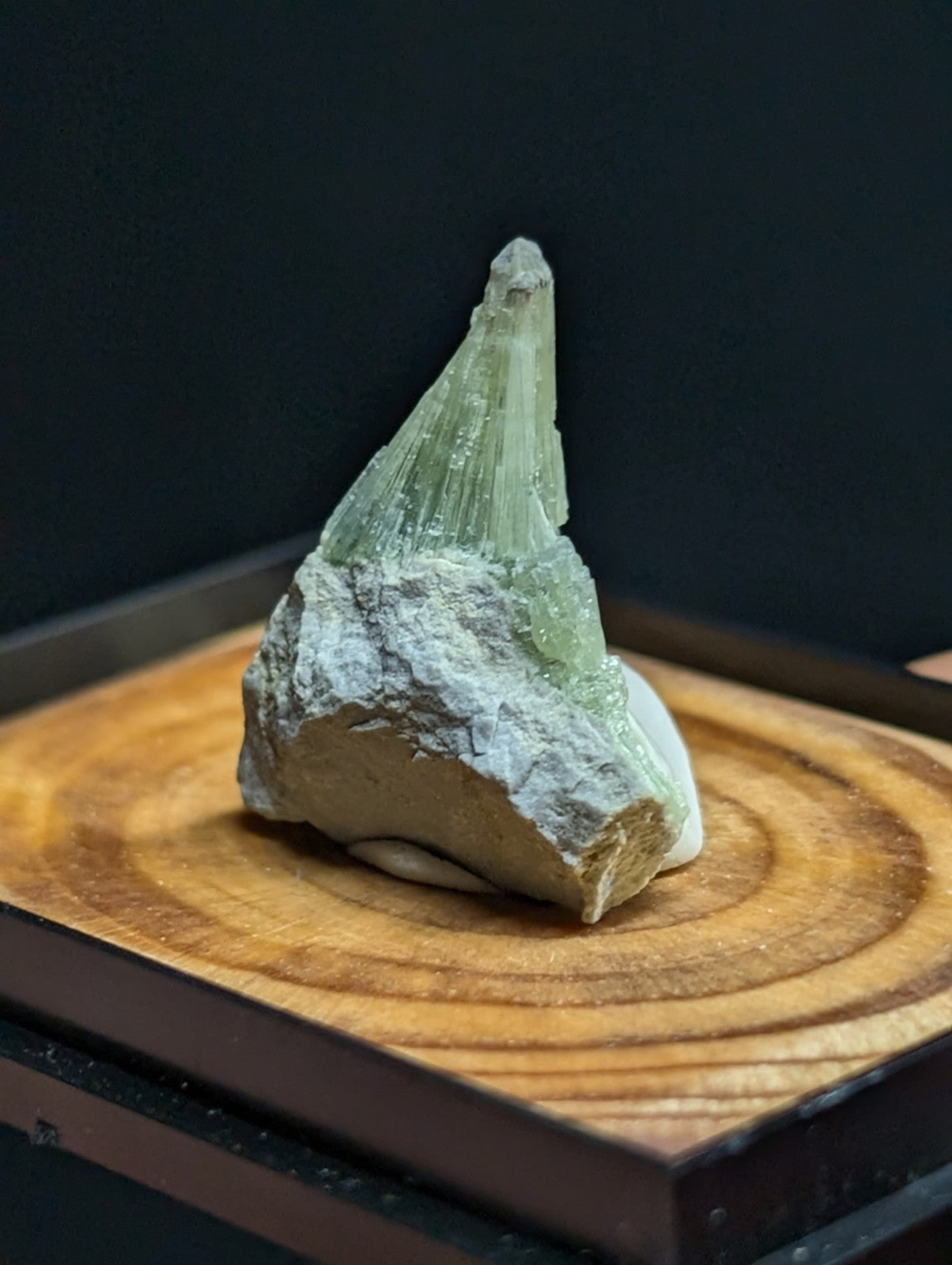 A translucent green crystal mineral specimen with a pointed, faceted tip emerging from a rough, light-colored rock base.