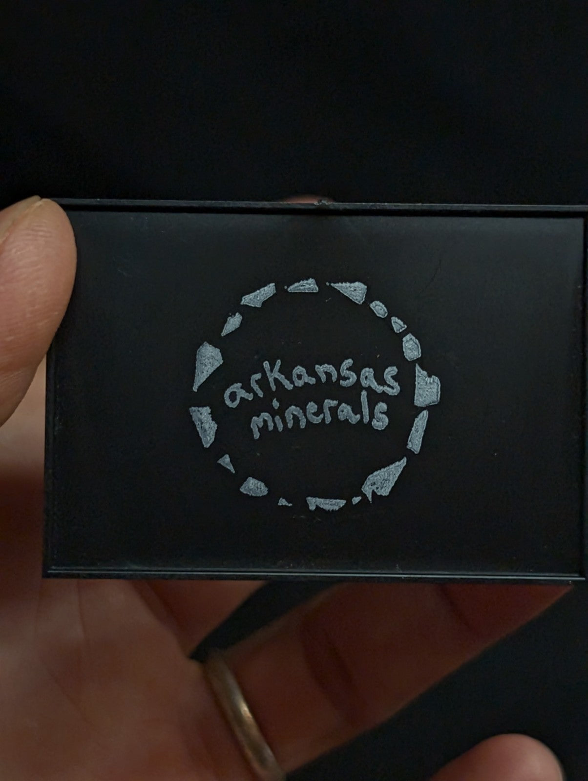 Black rectangular box with silver ’arkansas minerals’ logo encircled by a ring of stylized mineral fragments.