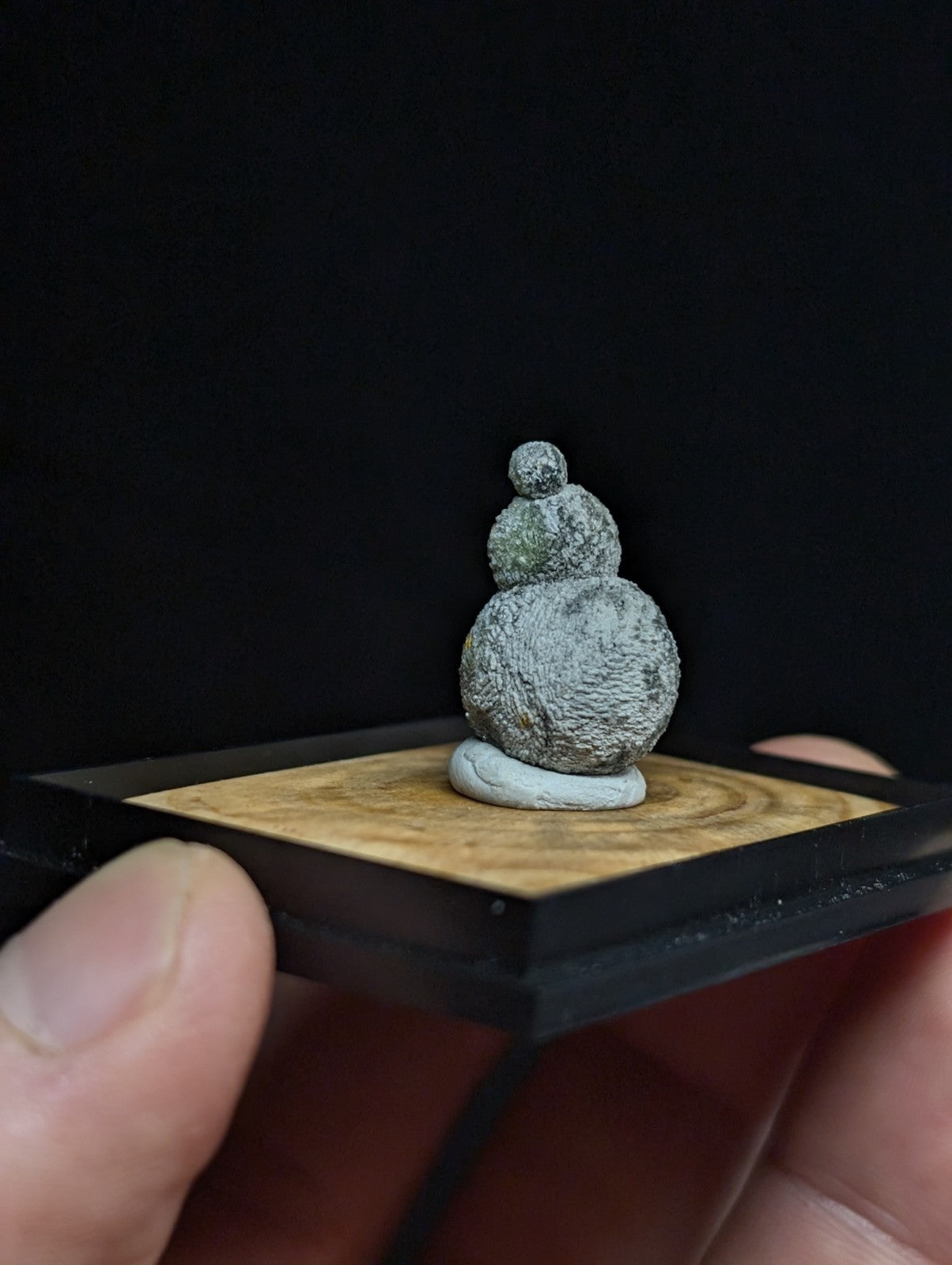 A miniature gray, textured stone sculpture resembling stacked gourds sits on a light wood base within a black frame.