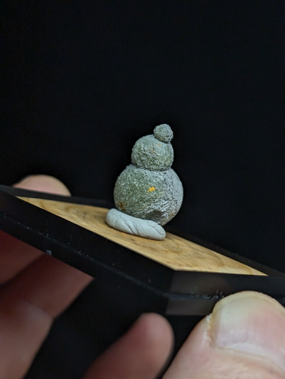 A miniature, textured green clay figurine resembling a seated snowman with a small yellow dot on its body and topped by a tiny round ball.