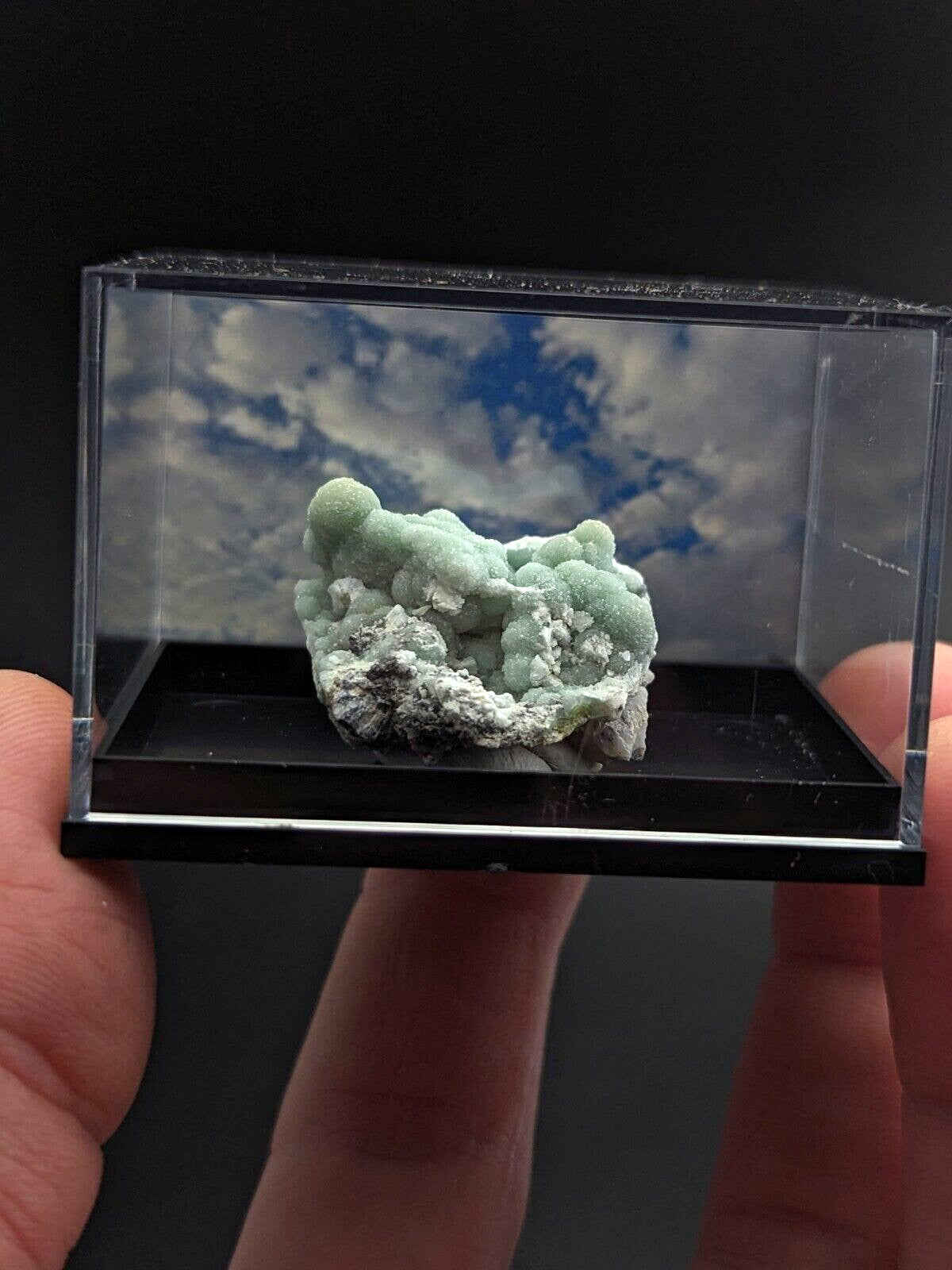A pale green, textured mineral specimen with white crystalline patches displayed in a clear acrylic display case.
