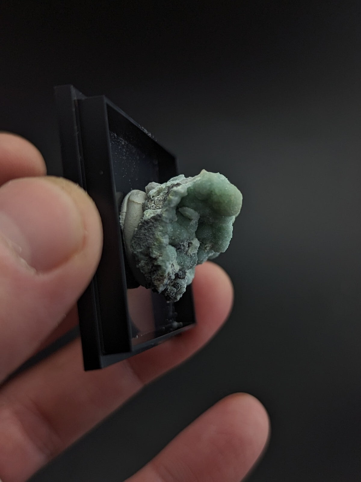 A pale green, textured mineral specimen with a rough, crystalline surface and darker grayish patches is displayed in a sleek black rectangular display case.