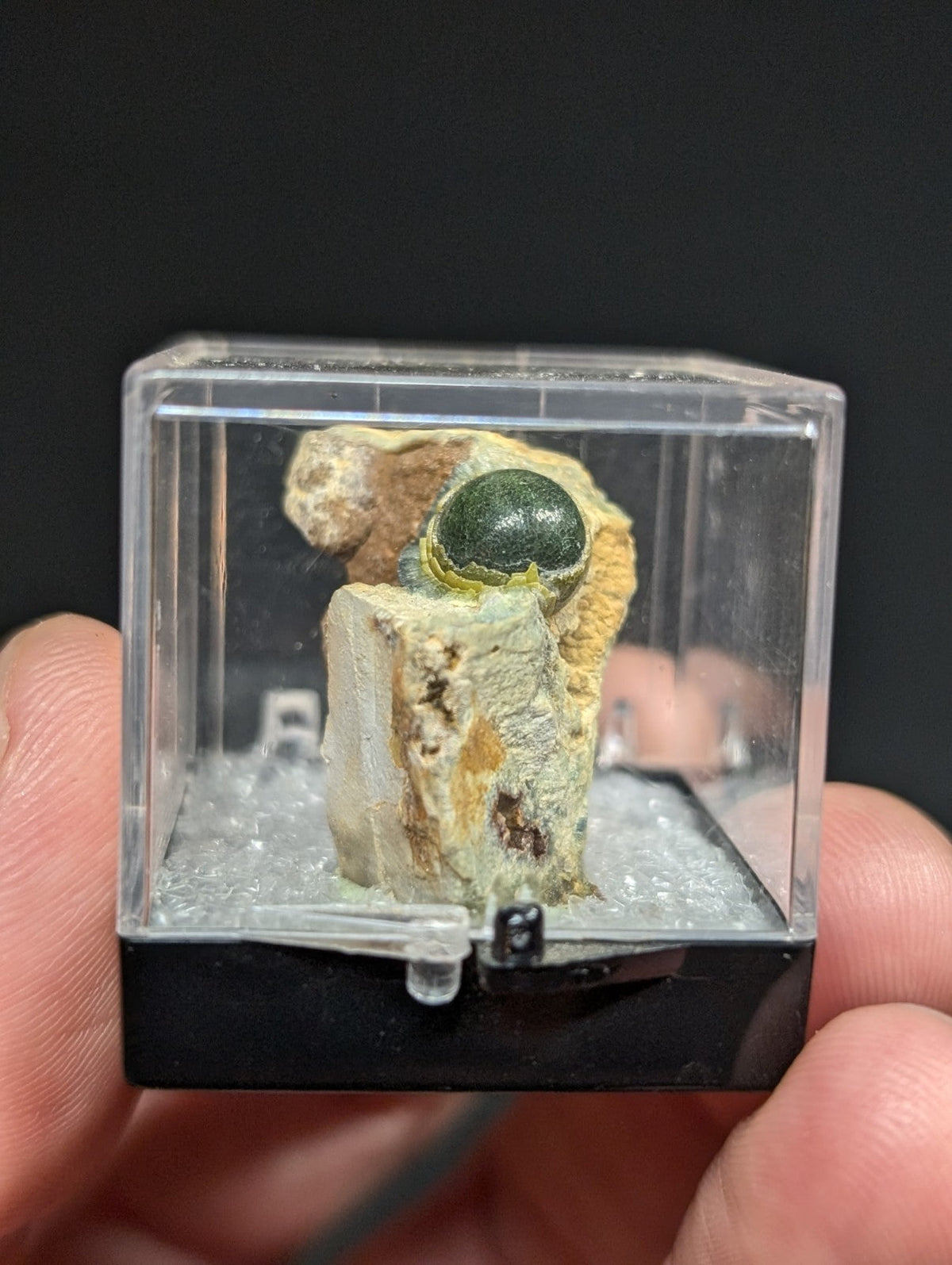 A polished green spherical mineral specimen nestled in a rocky matrix, encased within a clear acrylic display box with a black base.
