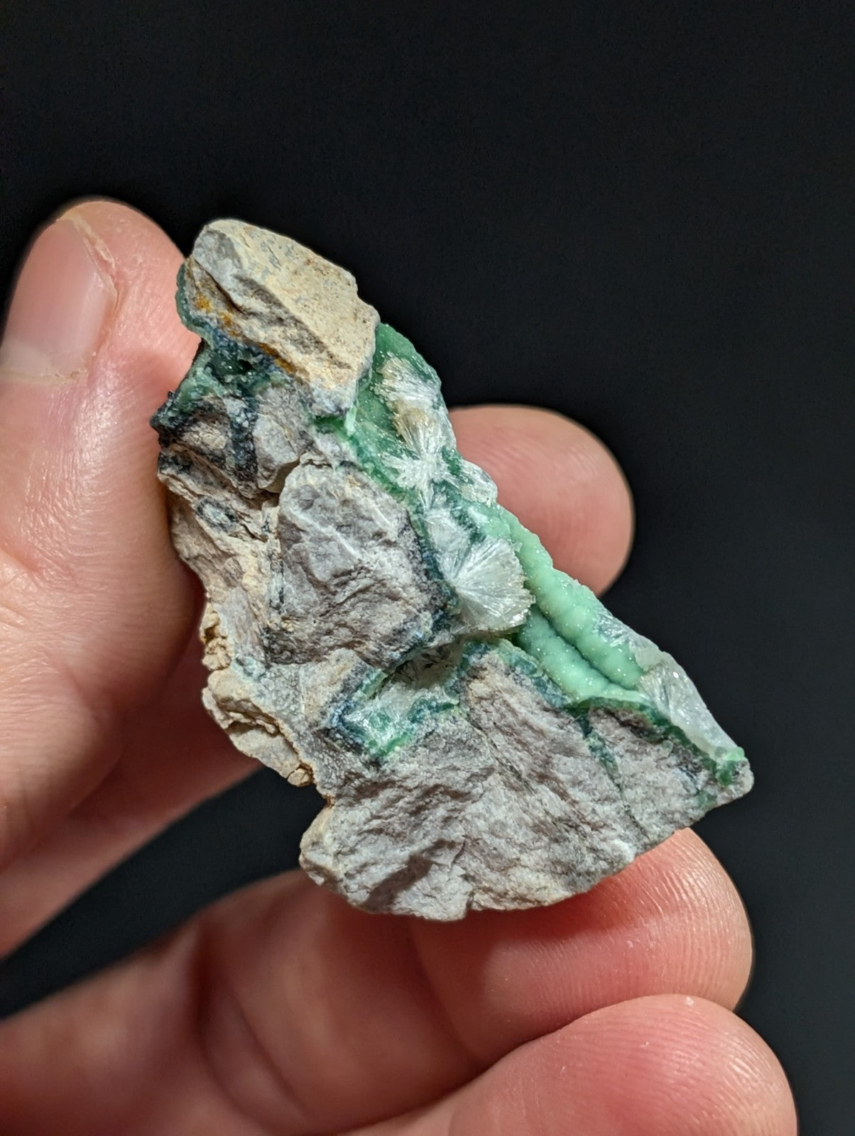 A hand holds a rough, irregularly shaped mineral specimen featuring vibrant green veining against a grayish matrix.