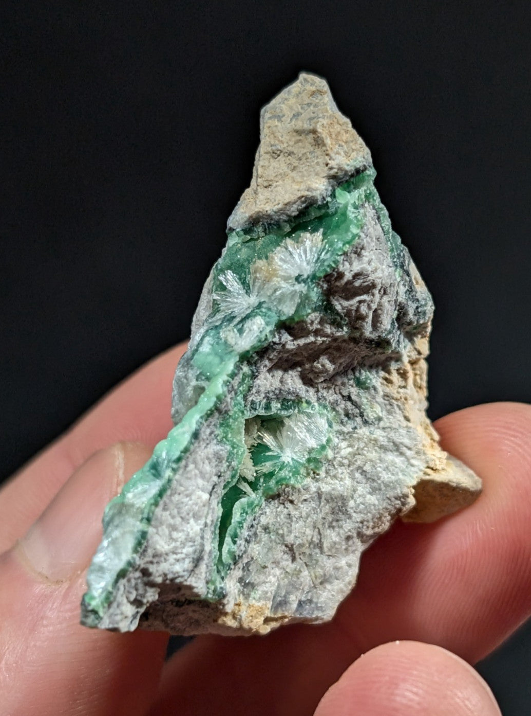 A hand holds a rough, irregularly shaped mineral specimen featuring vibrant green crystalline formations with delicate, feathery textures embedded in a grayish matrix.
