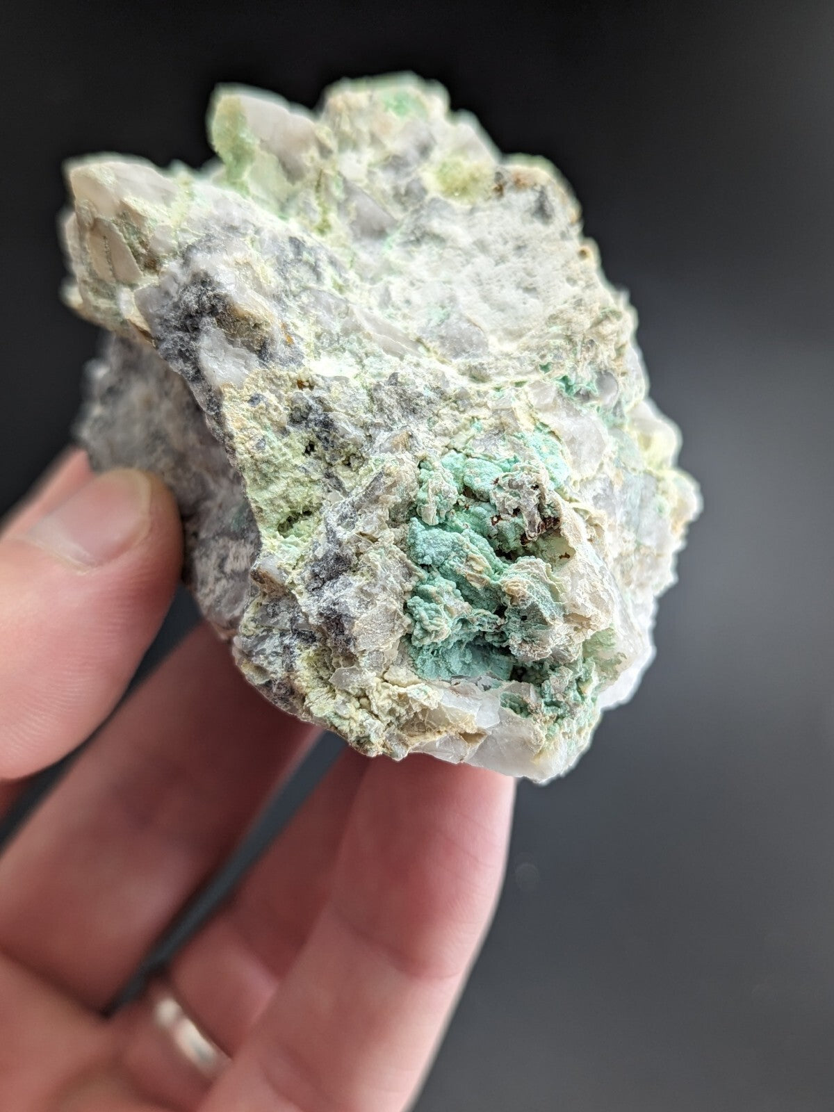 A hand holds a rough, irregularly shaped mineral specimen featuring patches of translucent white and vibrant green crystals embedded in a grayish matrix.
