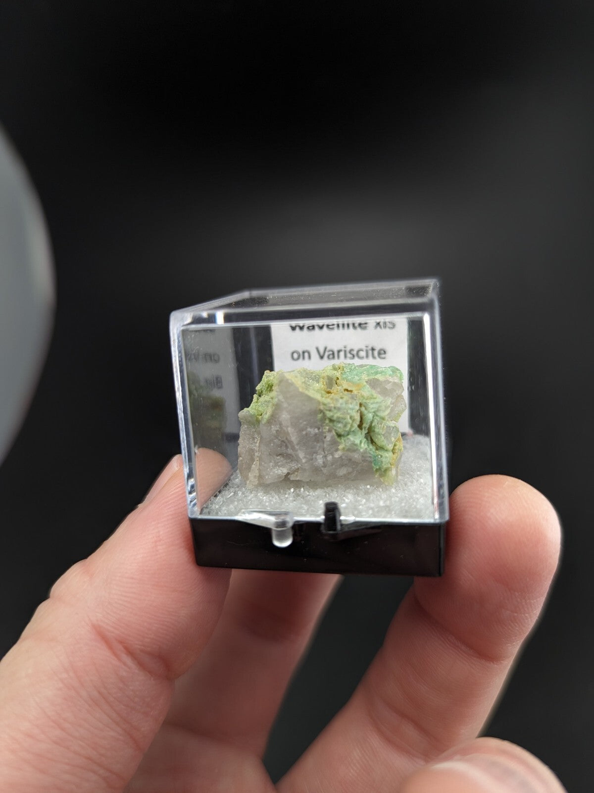 A green and white mineral specimen, waveite on variscite, displayed in a clear acrylic display case with a black base.