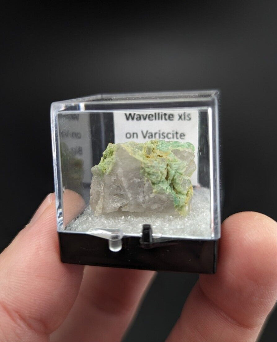 A translucent green and white mineral specimen, labeled ’wavellite xis on variscite,’ displayed in a clear acrylic display case with a black base.