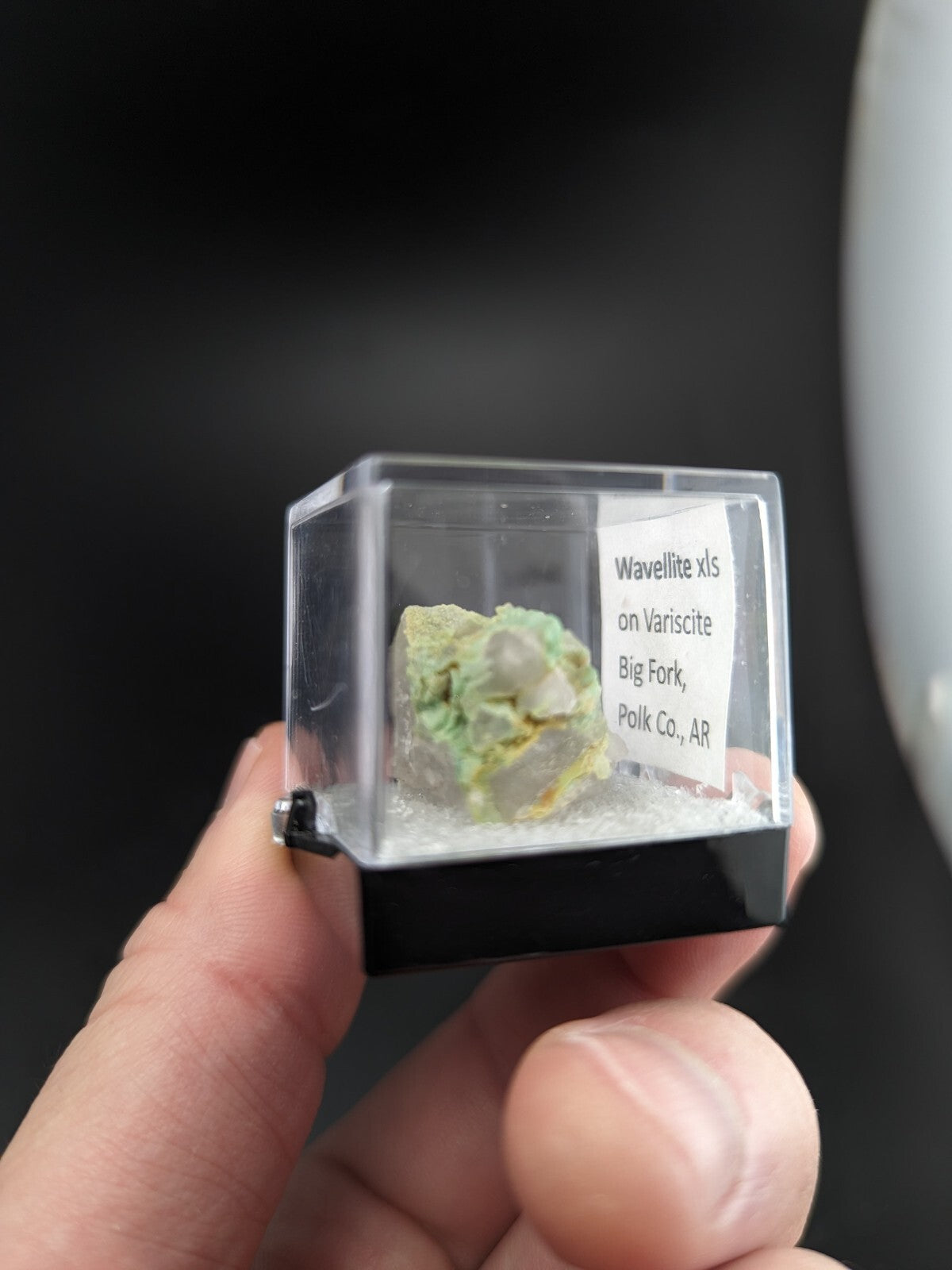 A green and yellow wavelite specimen encased in a clear plastic display box with a black base.