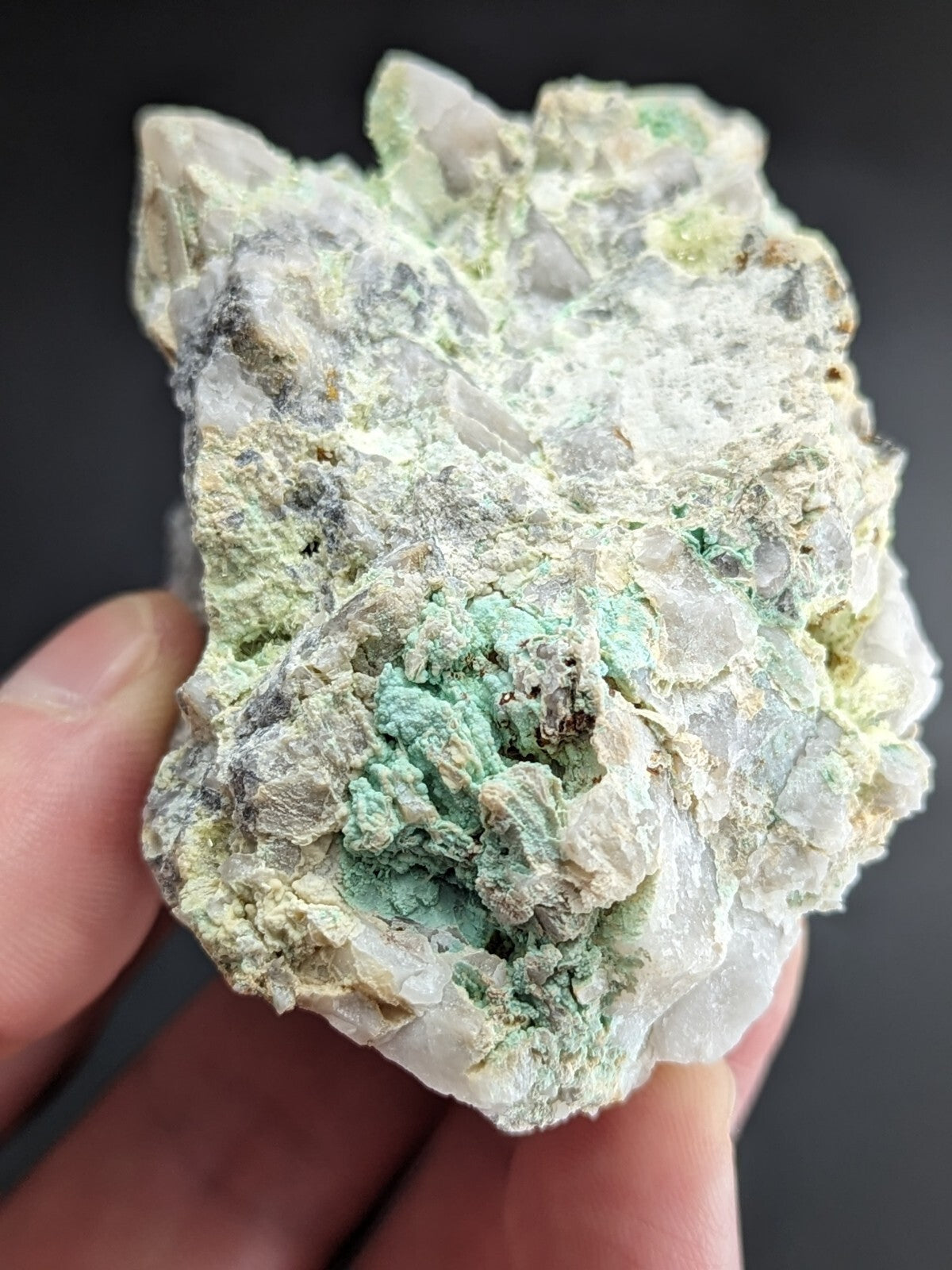 A hand holds a rough, irregularly shaped mineral specimen featuring translucent white crystals intermingled with patches of vibrant green and pale yellow hues.