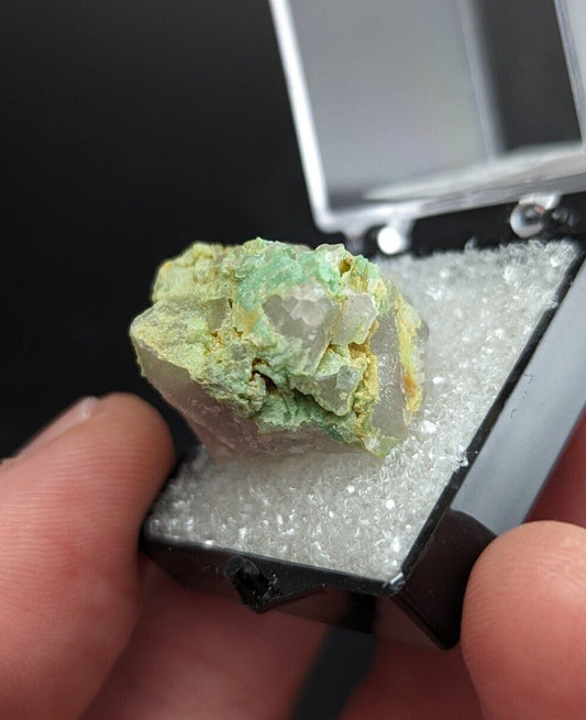 A green and white mineral specimen with crystalline formations, displayed in a black display case lined with crushed ice.