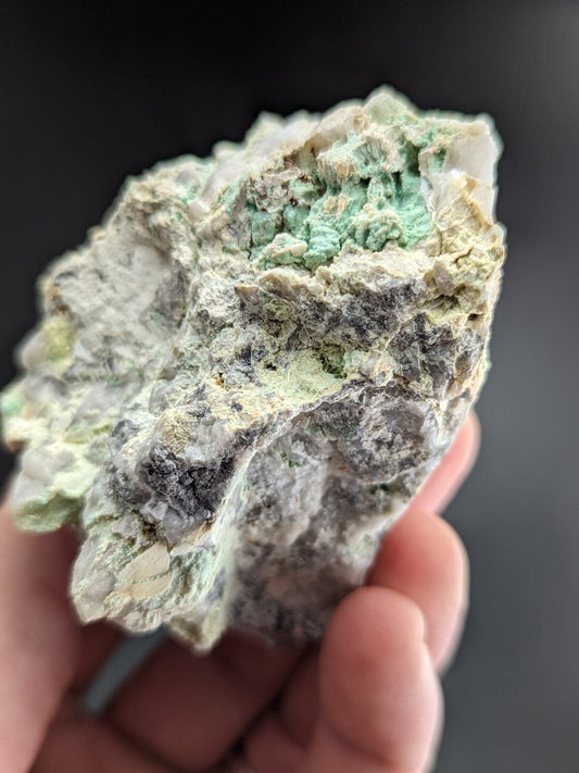 A rough, irregularly shaped mineral specimen with patches of vibrant green crystals embedded in a matrix of gray and white rock.