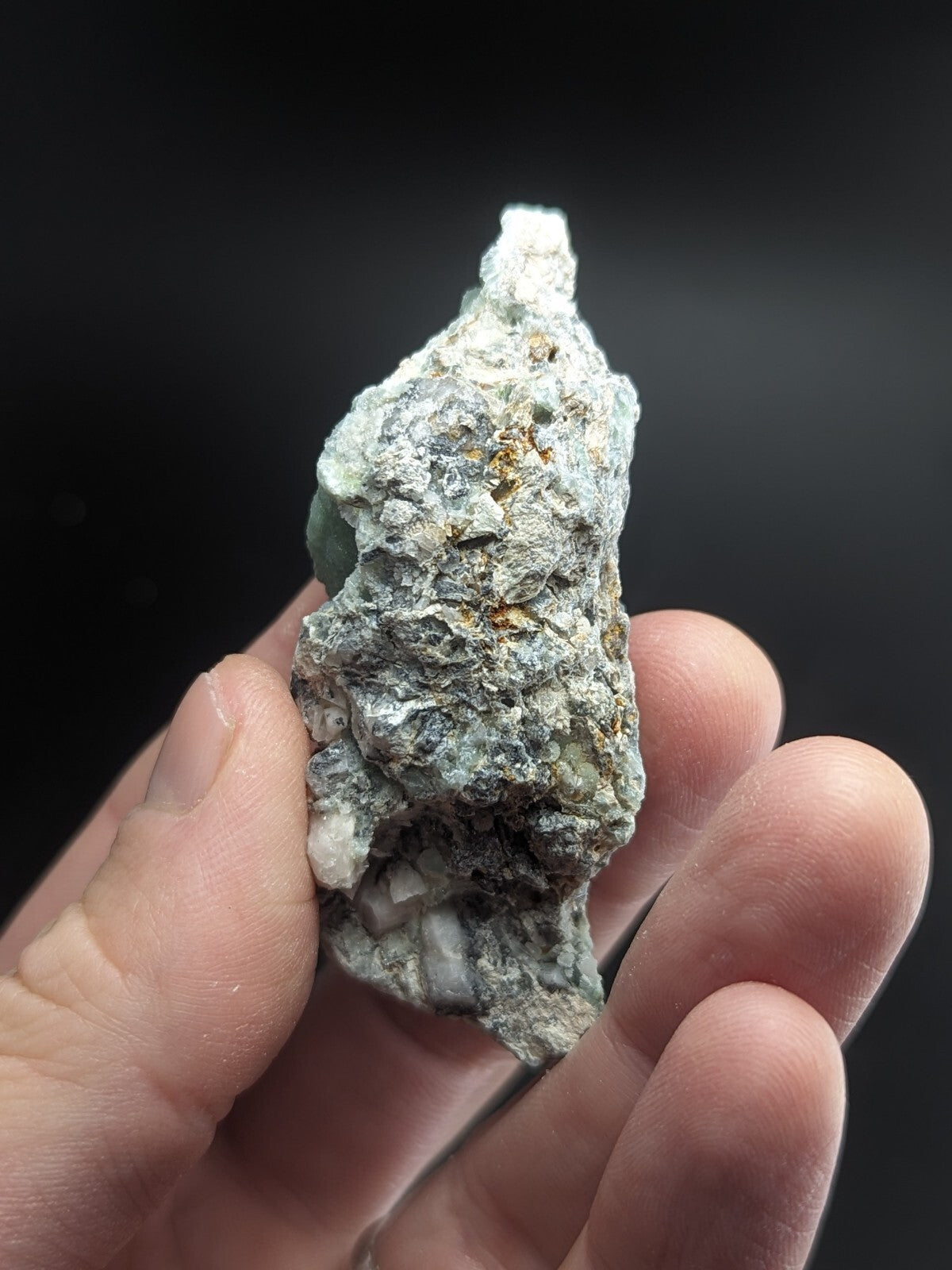 A rough, irregularly shaped mineral specimen with a mottled green and gray surface, featuring small white crystalline formations embedded in its rocky matrix.