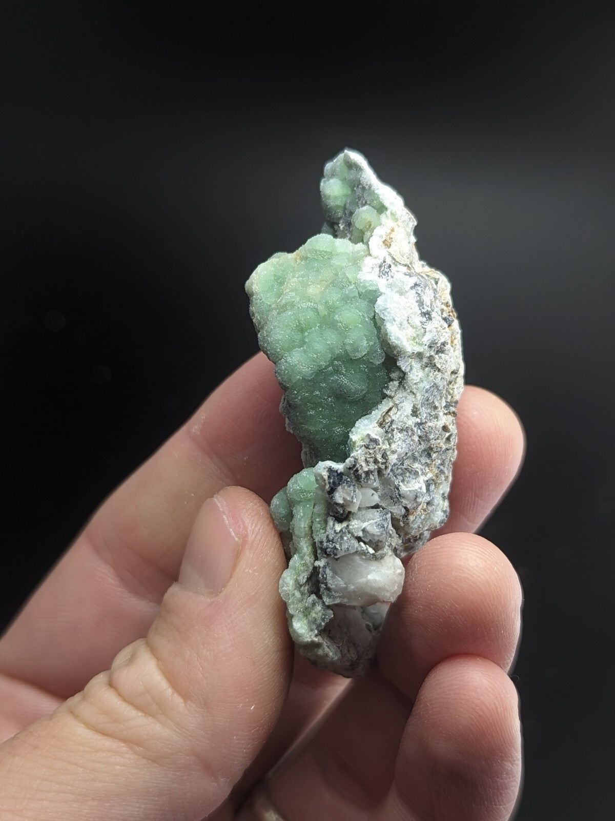 A hand holds a rough, pale green mineral specimen with crystalline textures and patches of white matrix.