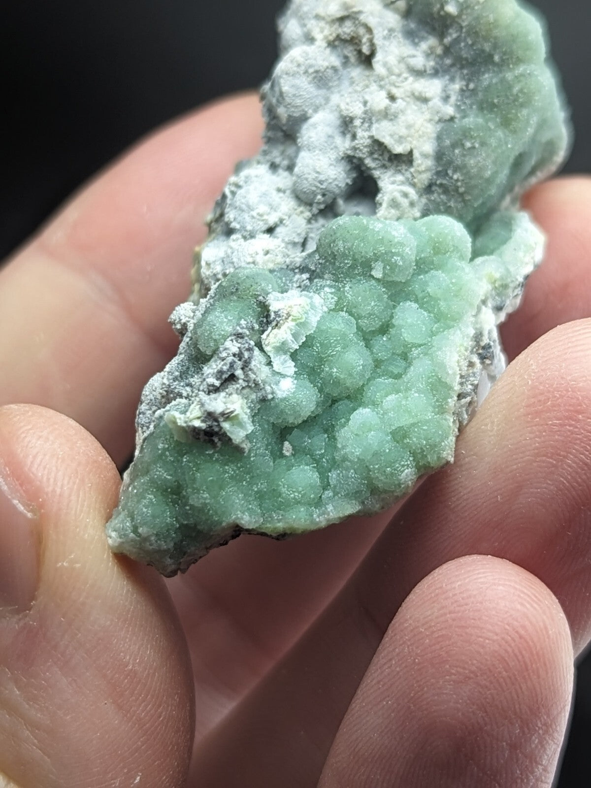A hand holds a green, bumpy mineral specimen with white crystalline patches.