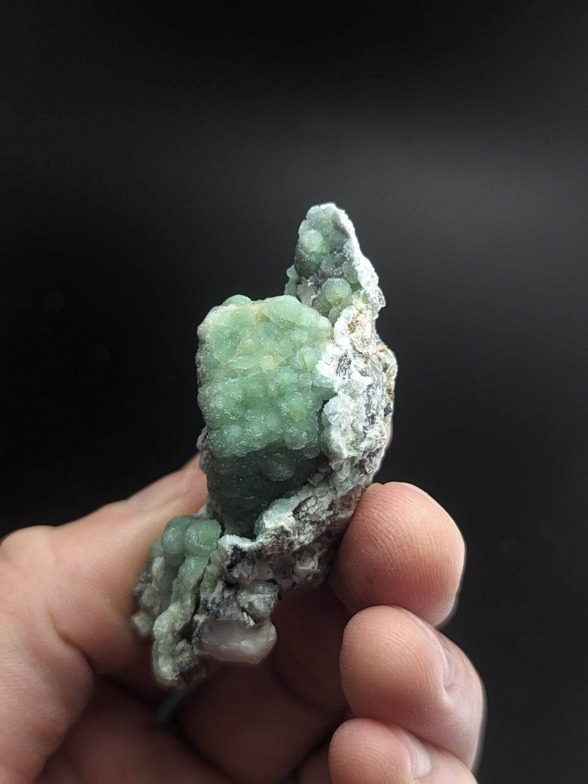 A hand holds a natural green mineral specimen with crystalline formations and a rough, earthy matrix.