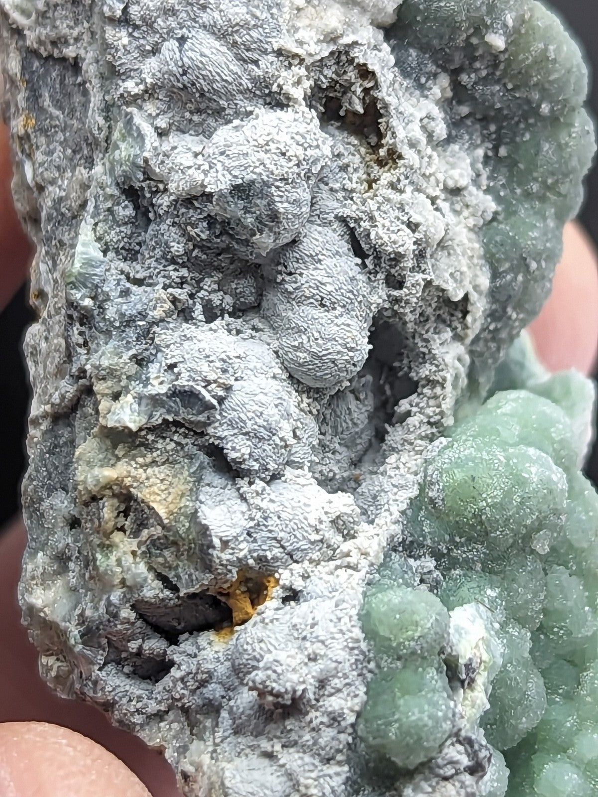 A textured mineral specimen featuring a mix of gray, white, and green crystalline formations with some yellowish accents.