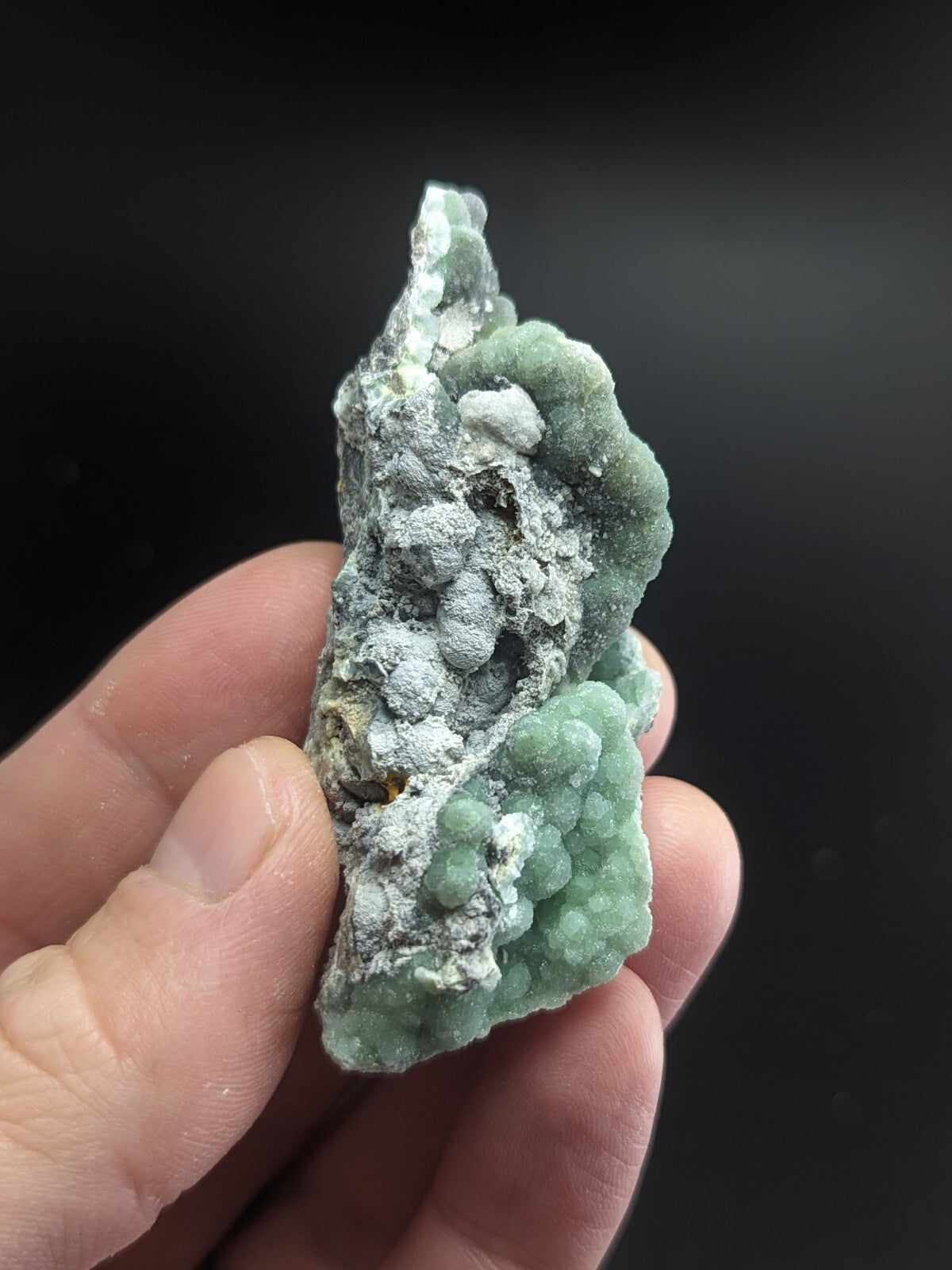 A hand holds a rough, irregularly shaped mineral specimen featuring clusters of light green crystals growing over a grayish matrix.
