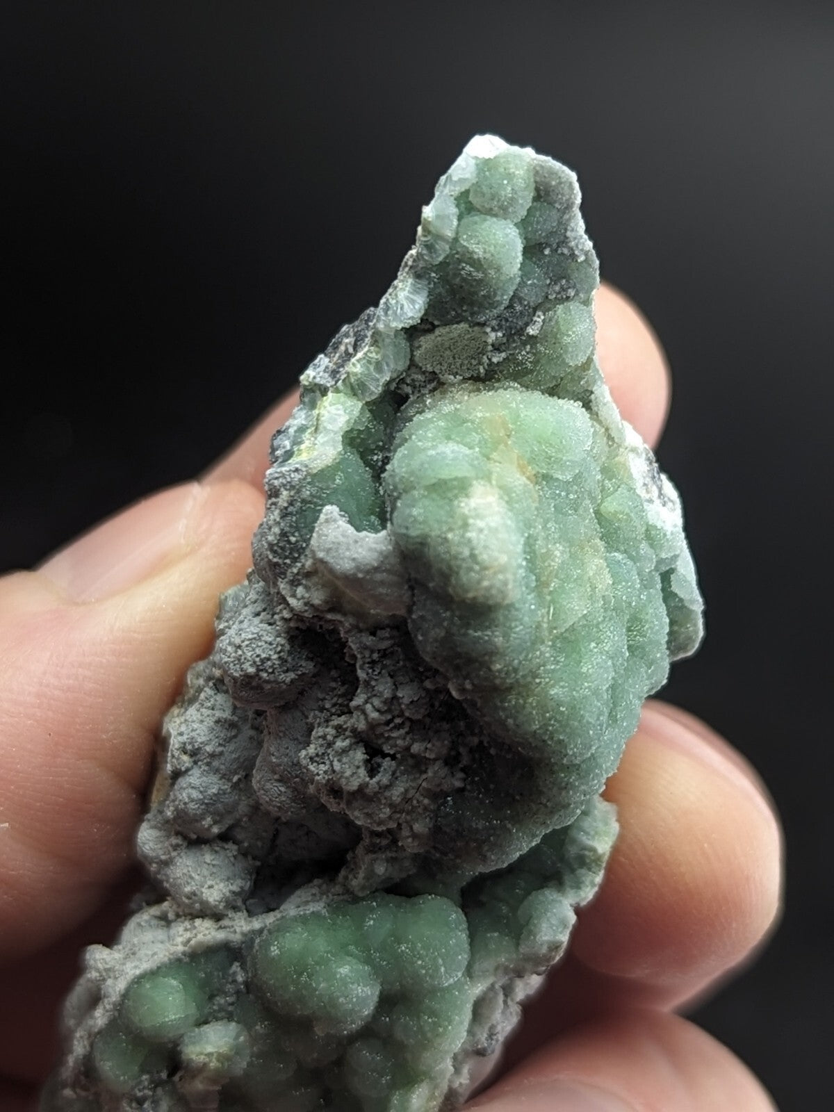 A hand holds a green, bumpy mineral specimen with translucent, clustered crystals growing from a grayish matrix.