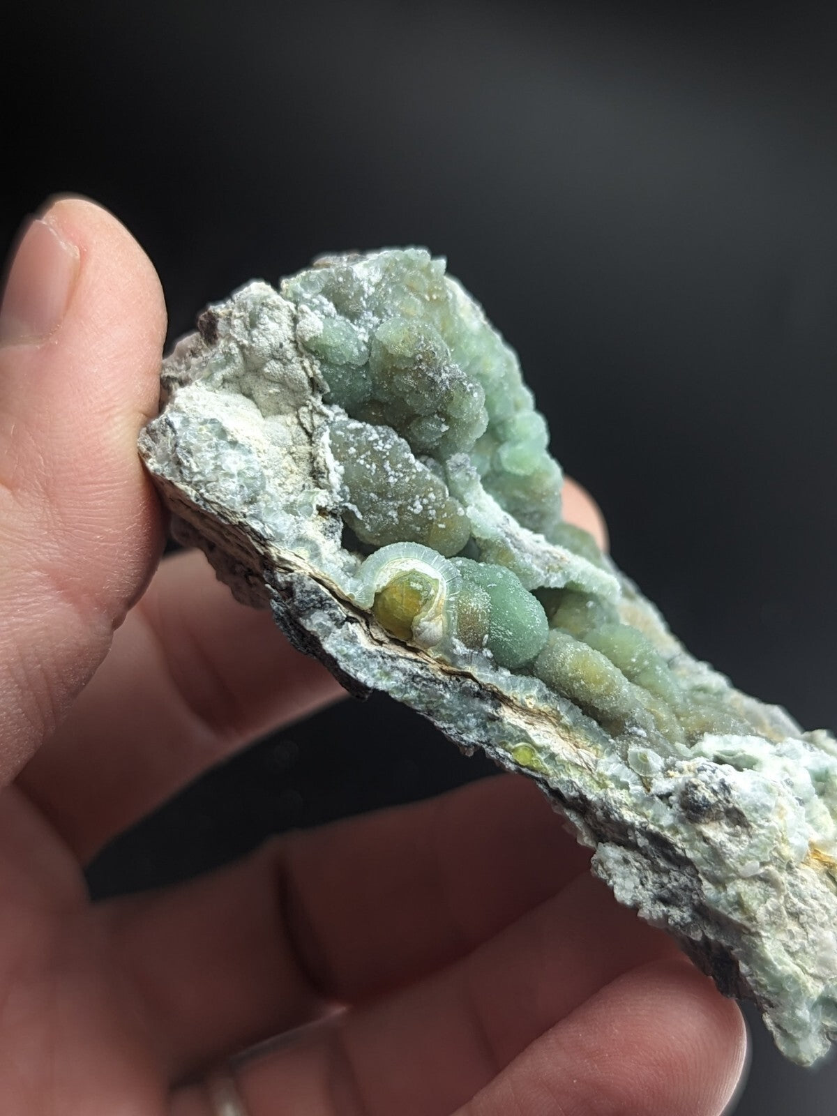 A hand holds a geode with translucent green crystals growing from a rough, grayish-white rock matrix.