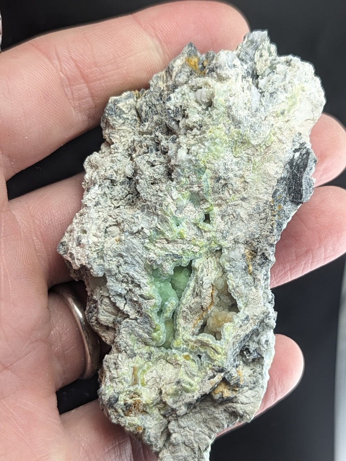 A rough, irregularly shaped mineral specimen with a mottled gray and green surface, featuring translucent patches of pale jade-green crystal embedded in its rocky matrix.