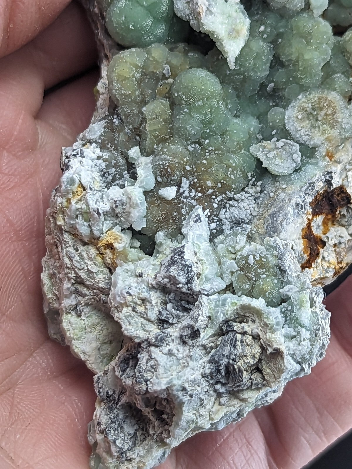 A hand holds a mineral specimen featuring clusters of translucent green crystals nestled within a matrix of white and gray rock, accented by patches of rust-colored material.