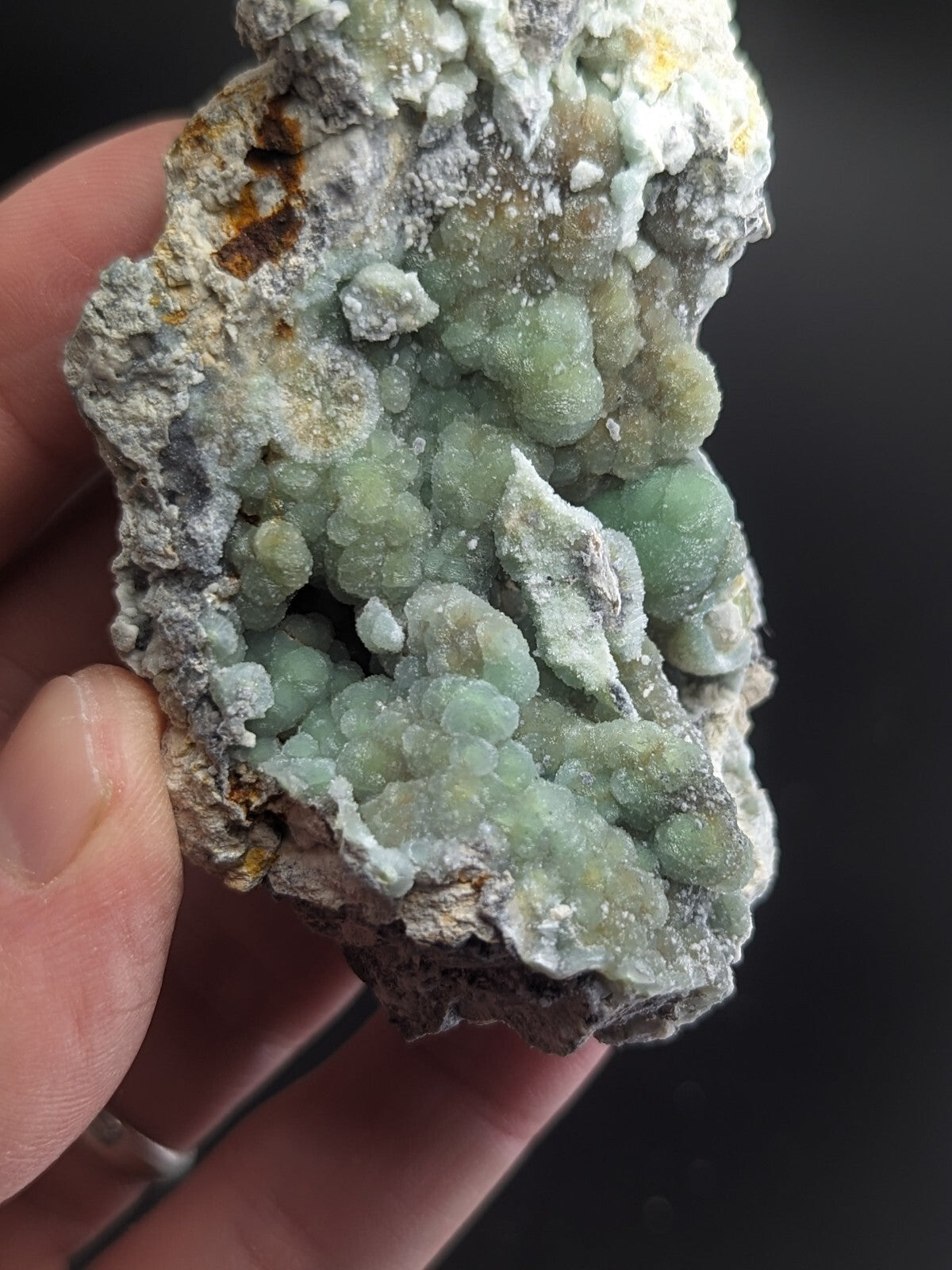 A hand holds a rough, irregularly shaped mineral specimen featuring clusters of translucent green crystals growing from a grayish matrix with patches of white and yellow.