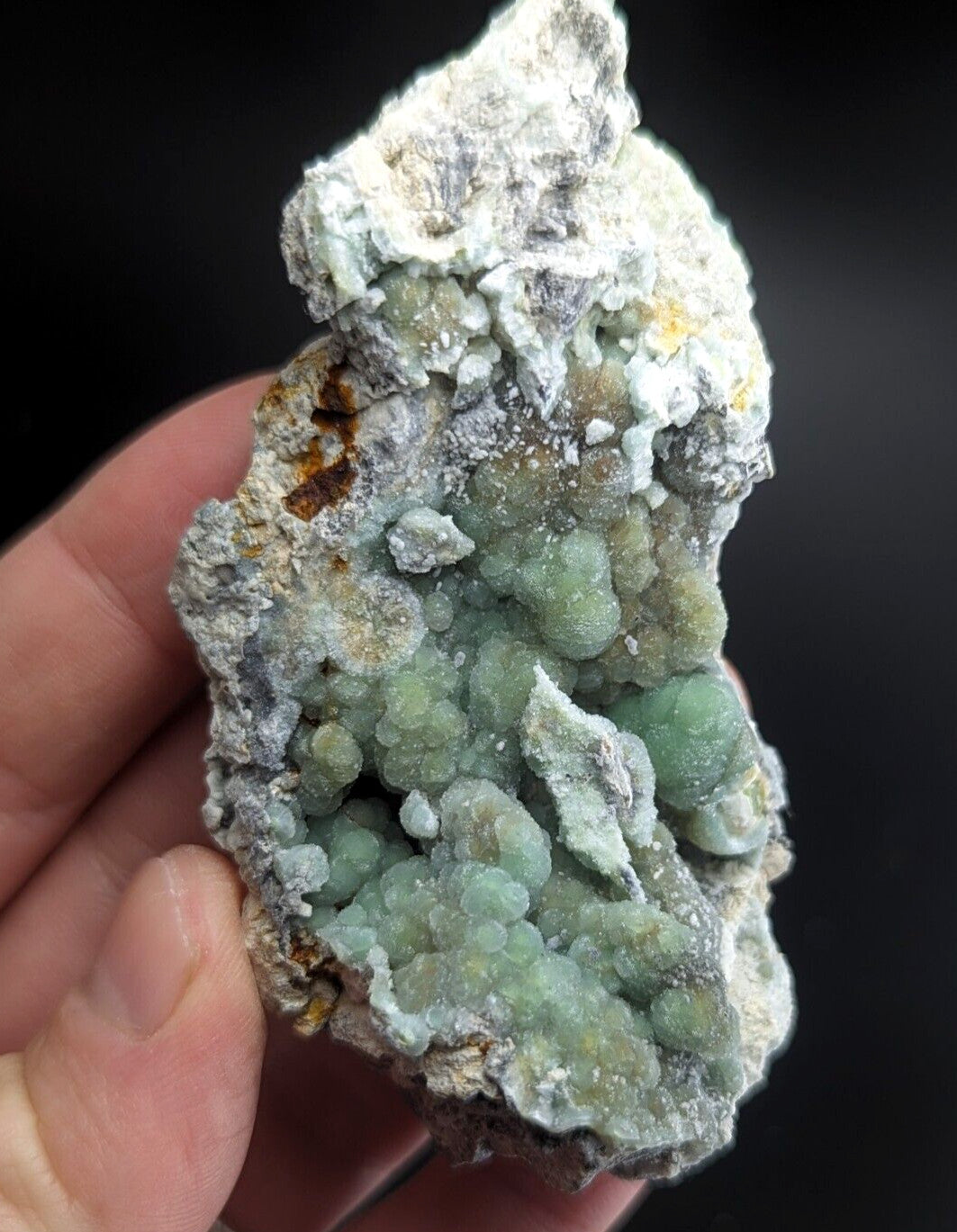 A hand holds a rough, irregularly shaped mineral specimen featuring clusters of translucent green crystals embedded in a matrix of white and gray rock.