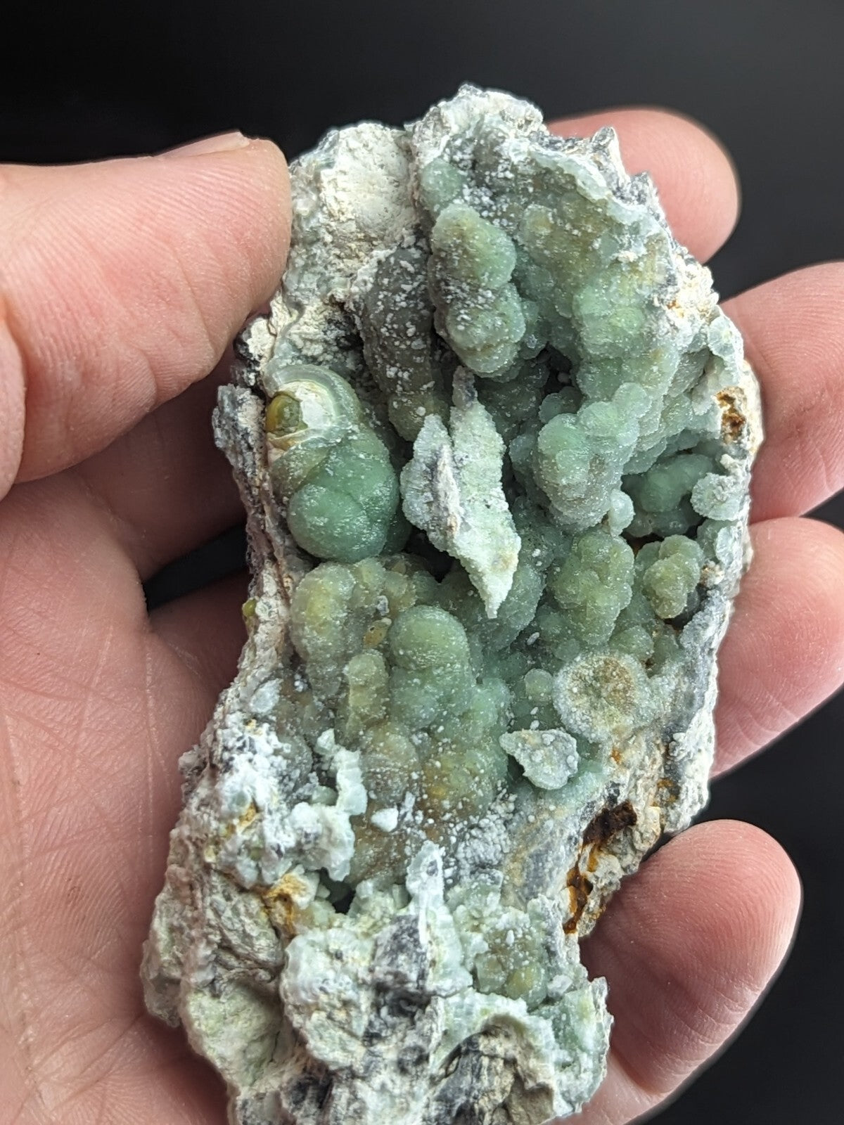 A hand holds a natural mineral specimen featuring clusters of translucent green crystals growing from a rough, grayish-white rock matrix.