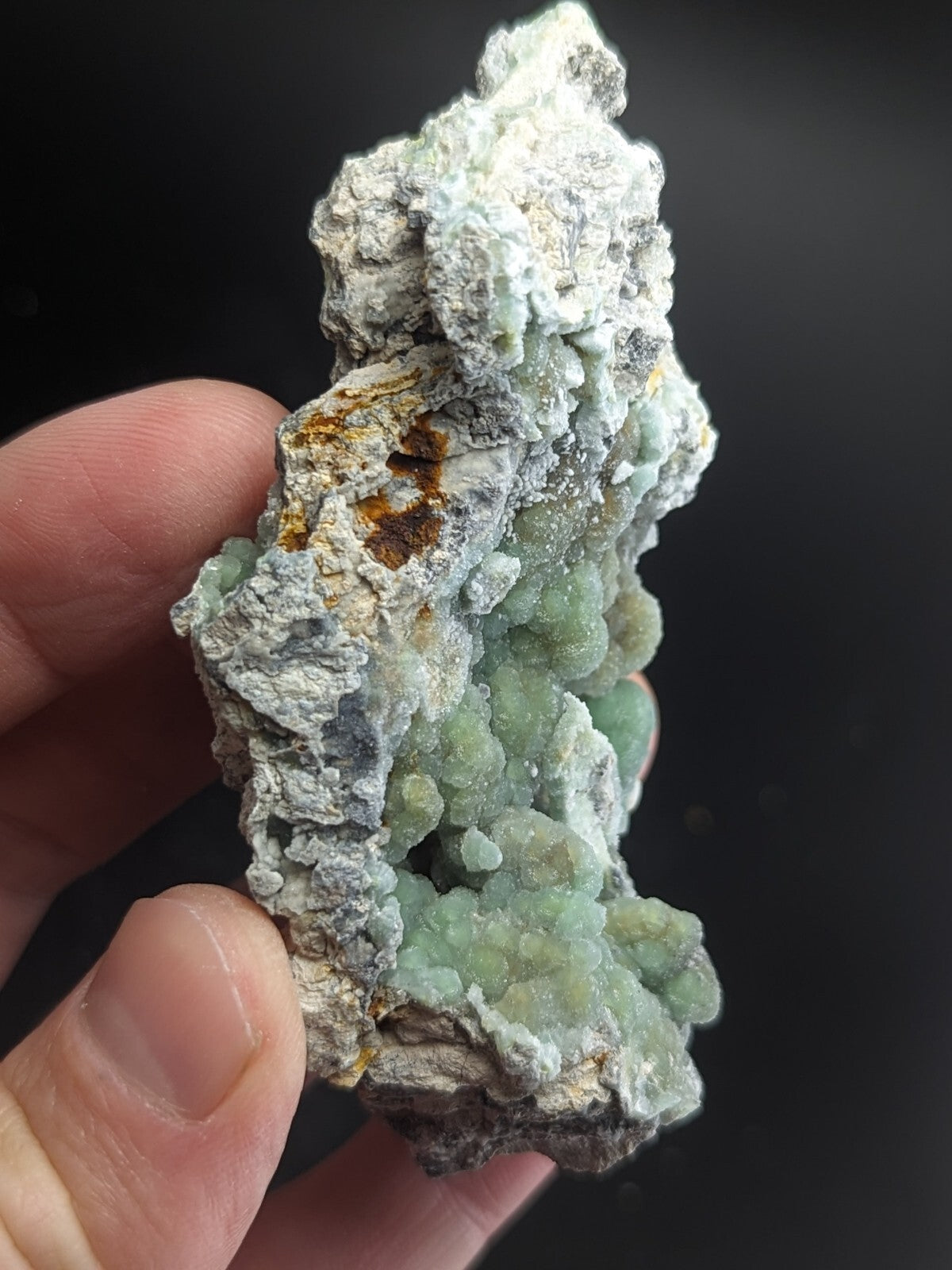 A hand holds a rough, irregularly shaped mineral specimen featuring clusters of translucent light green crystals growing from a matrix of grayish-white and brownish rock.